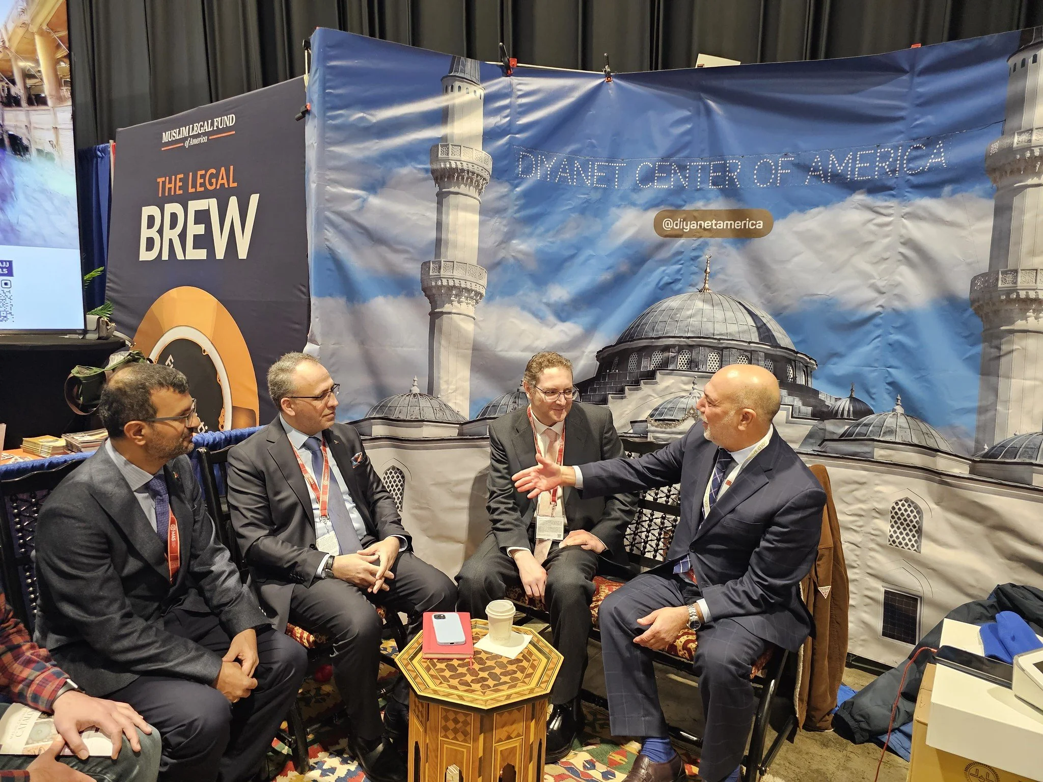 Five men in suits sitting and talking at an event booth with a backdrop featuring clouds and domed structures. The backdrop has text that reads 'Diyanet Center of America' and '@diyanetamerica.' One man is speaking with animated hand gestures.