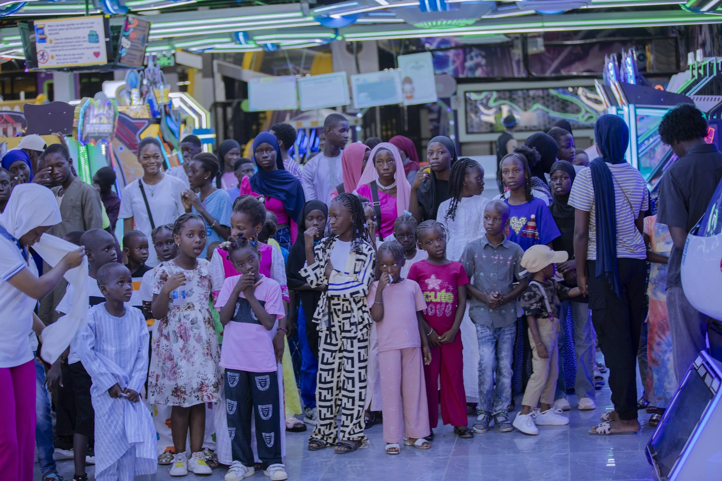A group of children and young teens gathered in an arcade or entertainment center with neon lights and gaming machines in the background.