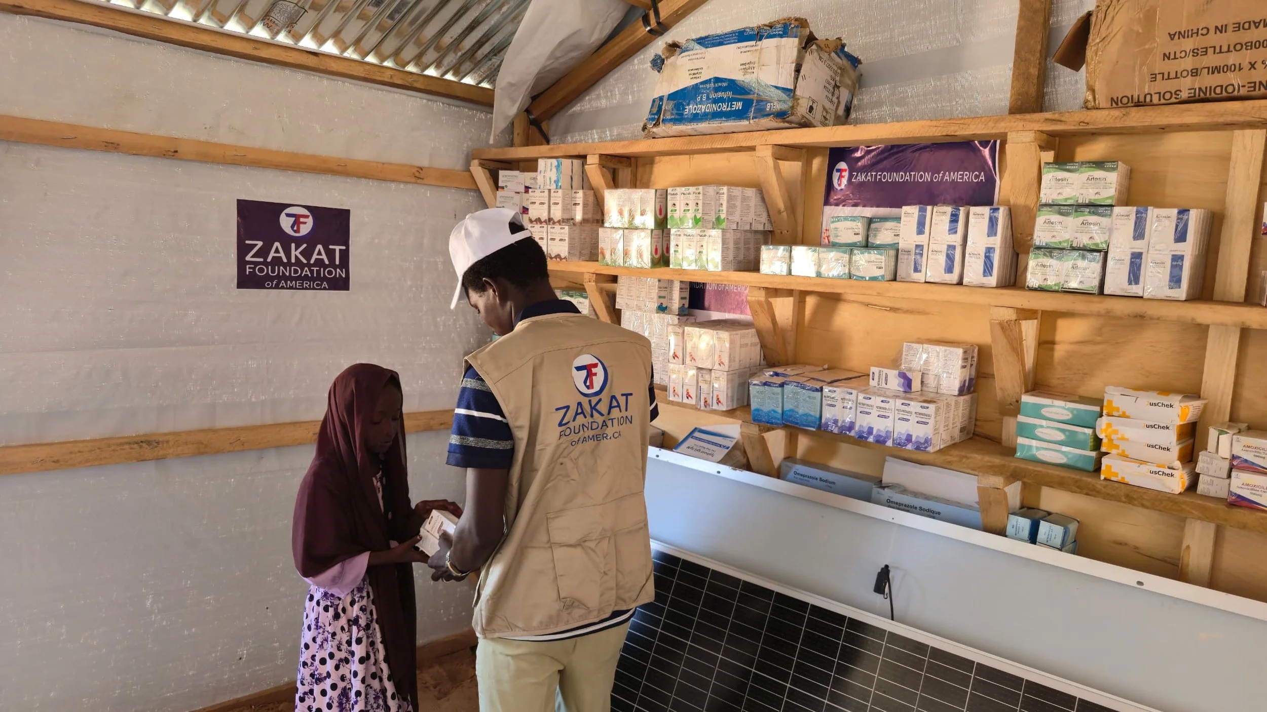 A person wearing a vest and a white cap from the Zakat Foundation of America is assisting a young girl in a modest dress inside a small room with shelves filled with medical supplies and medicines.