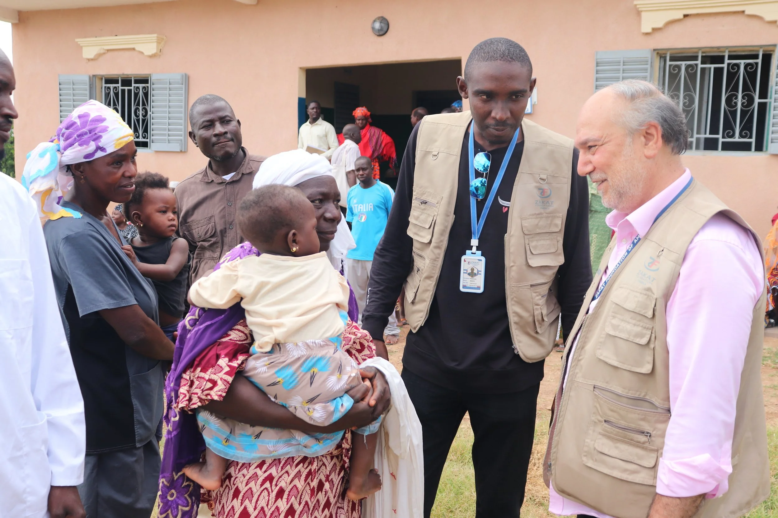 A group of people, including women and children, gather outside a building. An older woman is holding a young child and talking to two men wearing beige vests, one of whom has sunglasses hanging from his shirt. The background shows more people and a peach-colored building with windows.