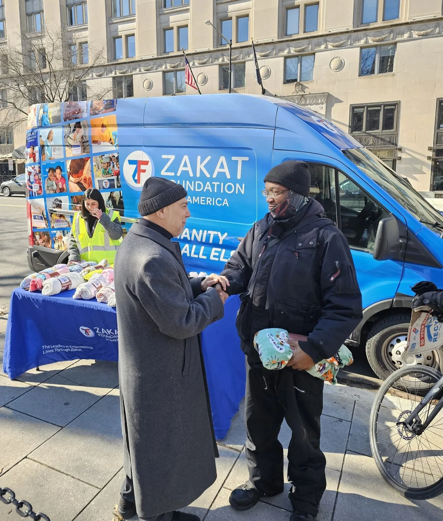 Two men shaking hands in front of a Zakat Foundation of America van at an outdoor charity event, with a table of knitted items and a woman in a yellow vest in the background.