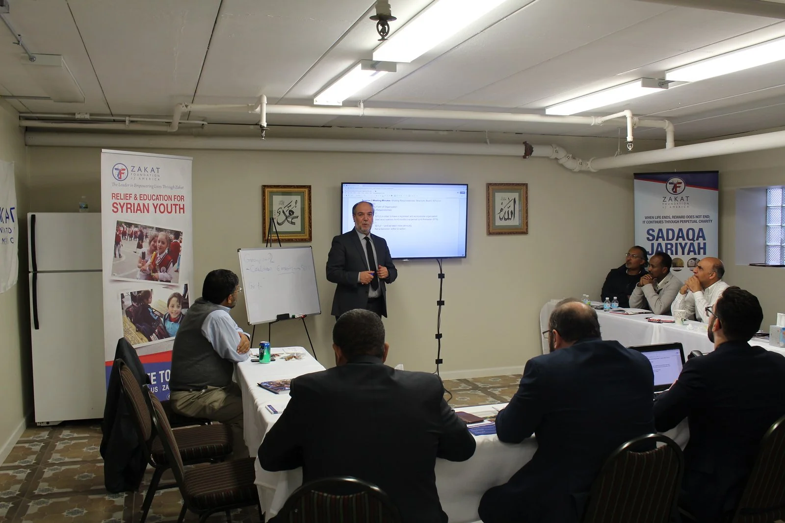 Halil Demir in a suit giving a presentation in a conference room with people seated at tables. There are banners on both sides promoting relief efforts for Syrian youth and a TV screen behind the presenter.