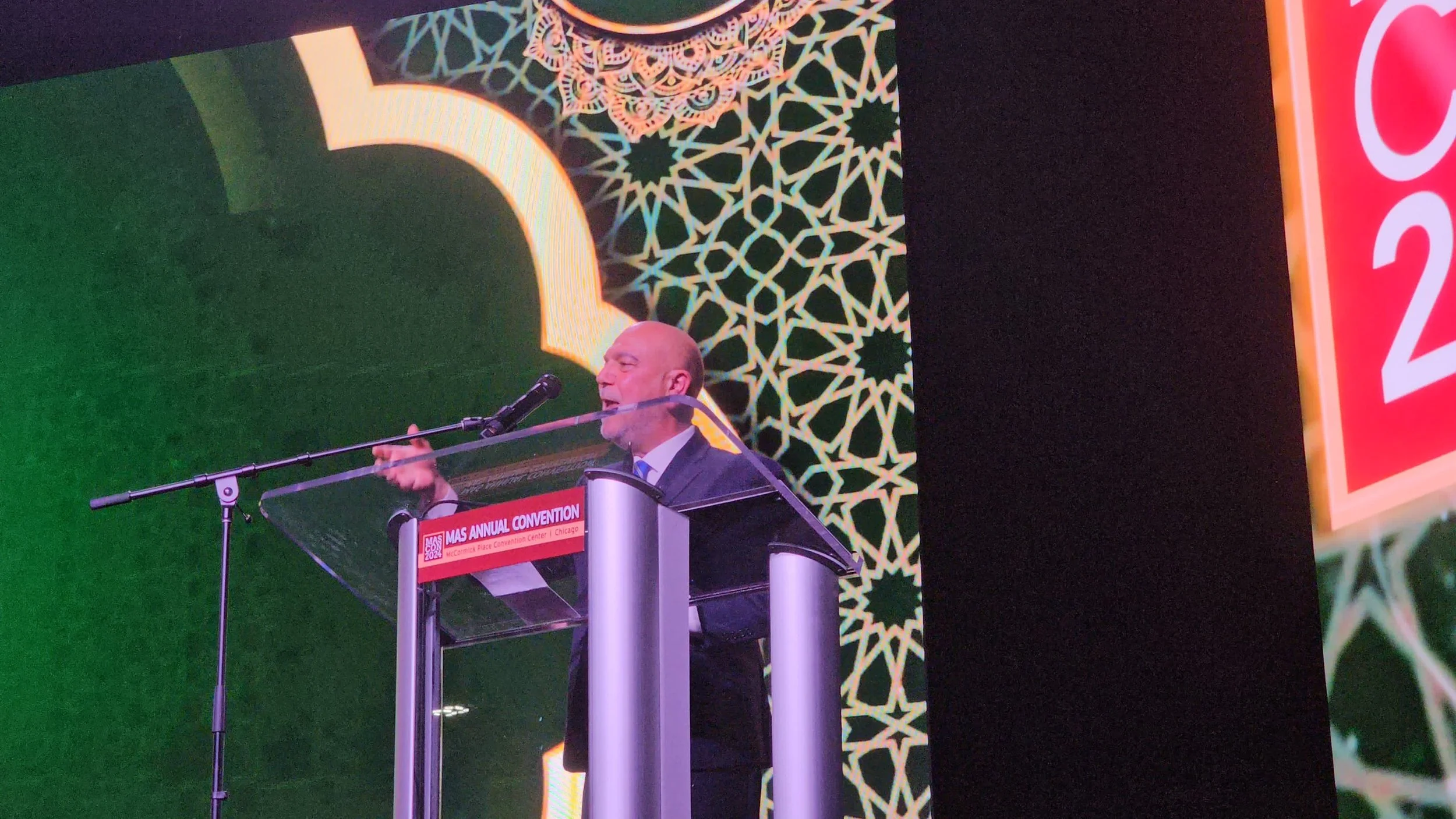 Halil Demir speaking at a podium during the MAS Annual Convention, with a large digital screen featuring green, black, and intricate gold geometric patterns in the background.