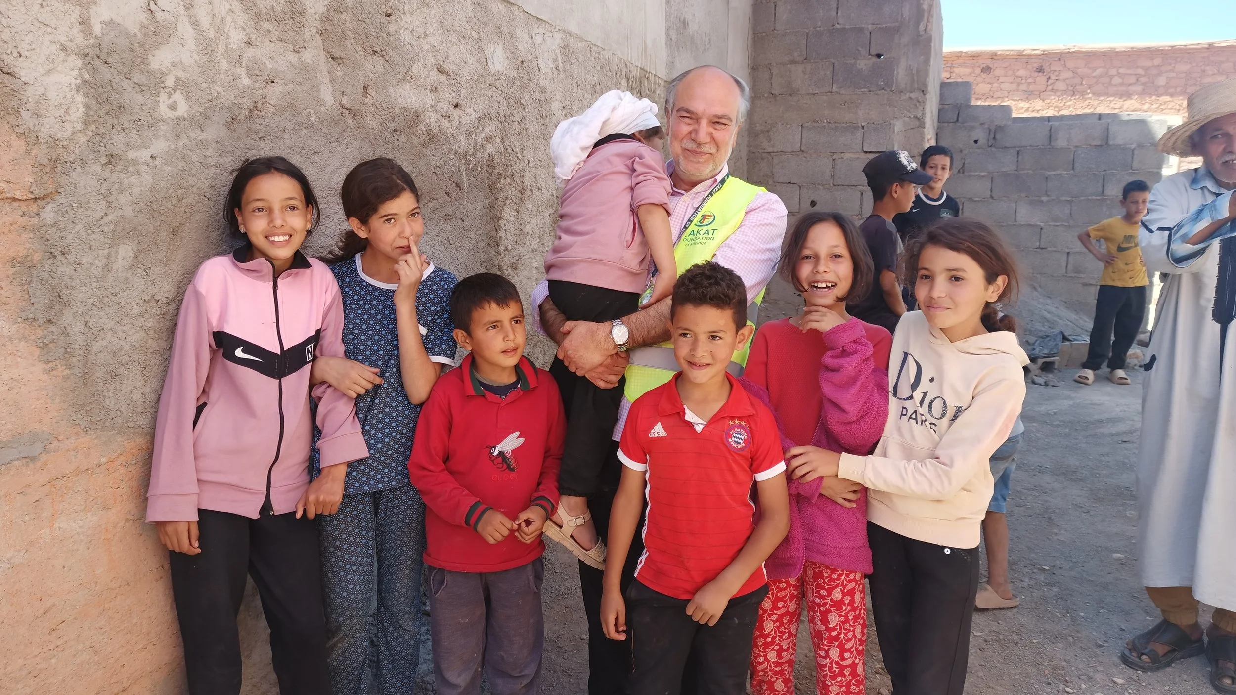 Halil Demir  wearing a yellow vest is holding a young girl on his shoulders, surrounded by smiling children of various ages in front of a rough wall, with other children and adults in the background.