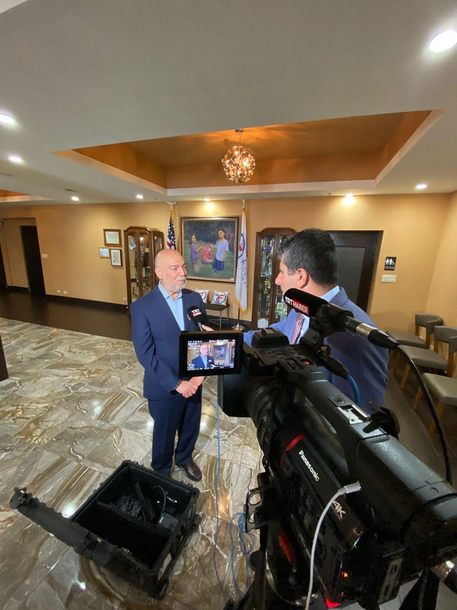 Halil Demir in a blue suit being interviewed by a news reporter in a room with warm-colored walls, flags, and a painting in the background.