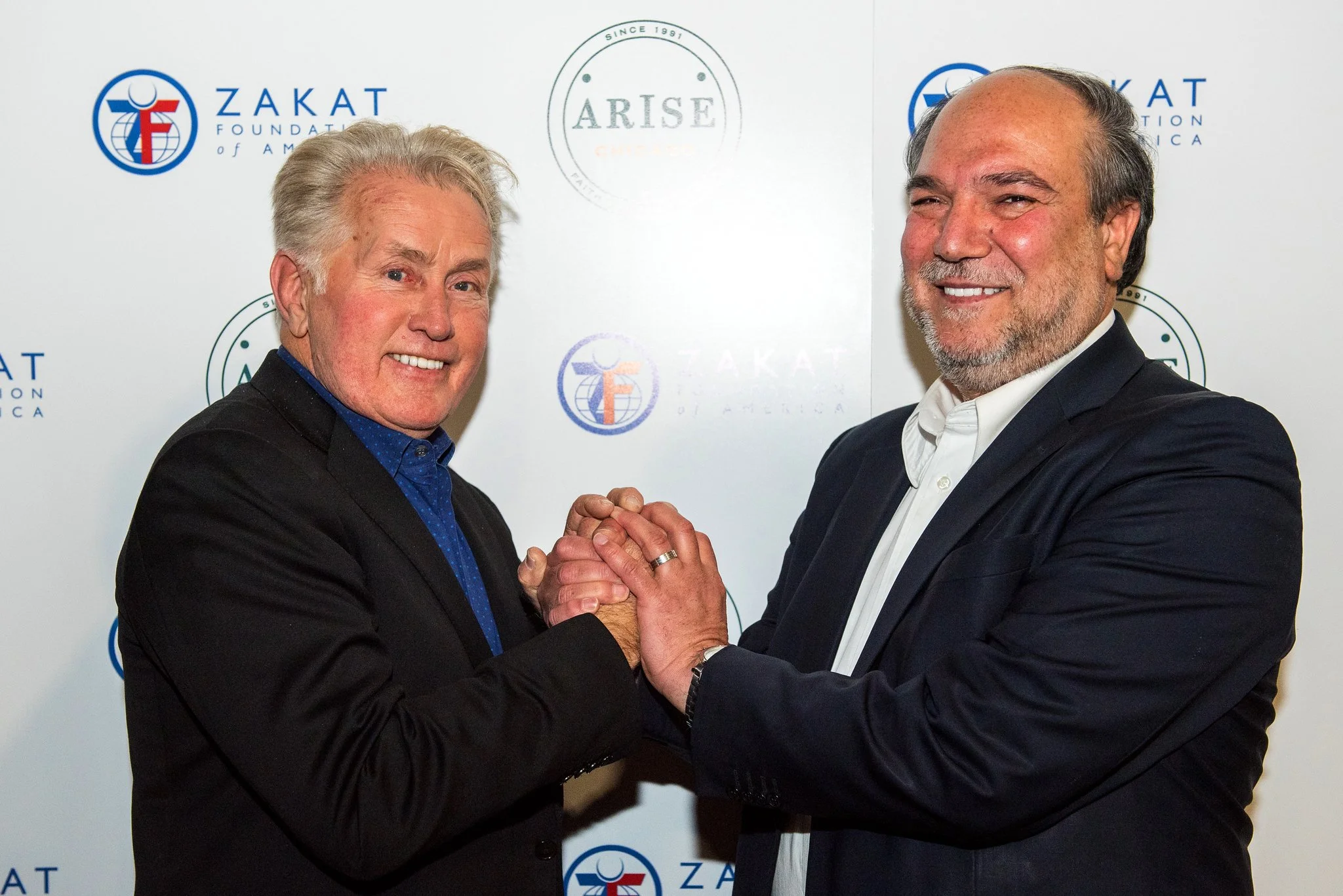 Halil Demir and Sheen in suits shaking hands and smiling, standing in front of a white backdrop with logos and text, including 'Zakat Foundation of America' and 'ARISE'.