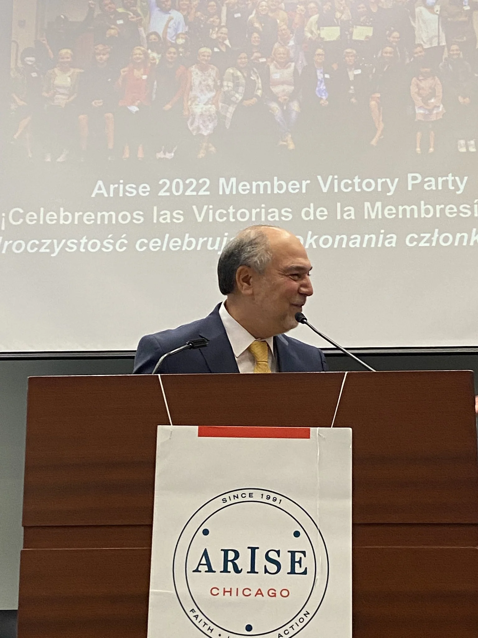 Halil Demir in a suit with a yellow tie standing behind a podium with an ARISE Chicago banner, speaking into a microphone at a conference or event, with a presentation slide in the background showing a group photo.