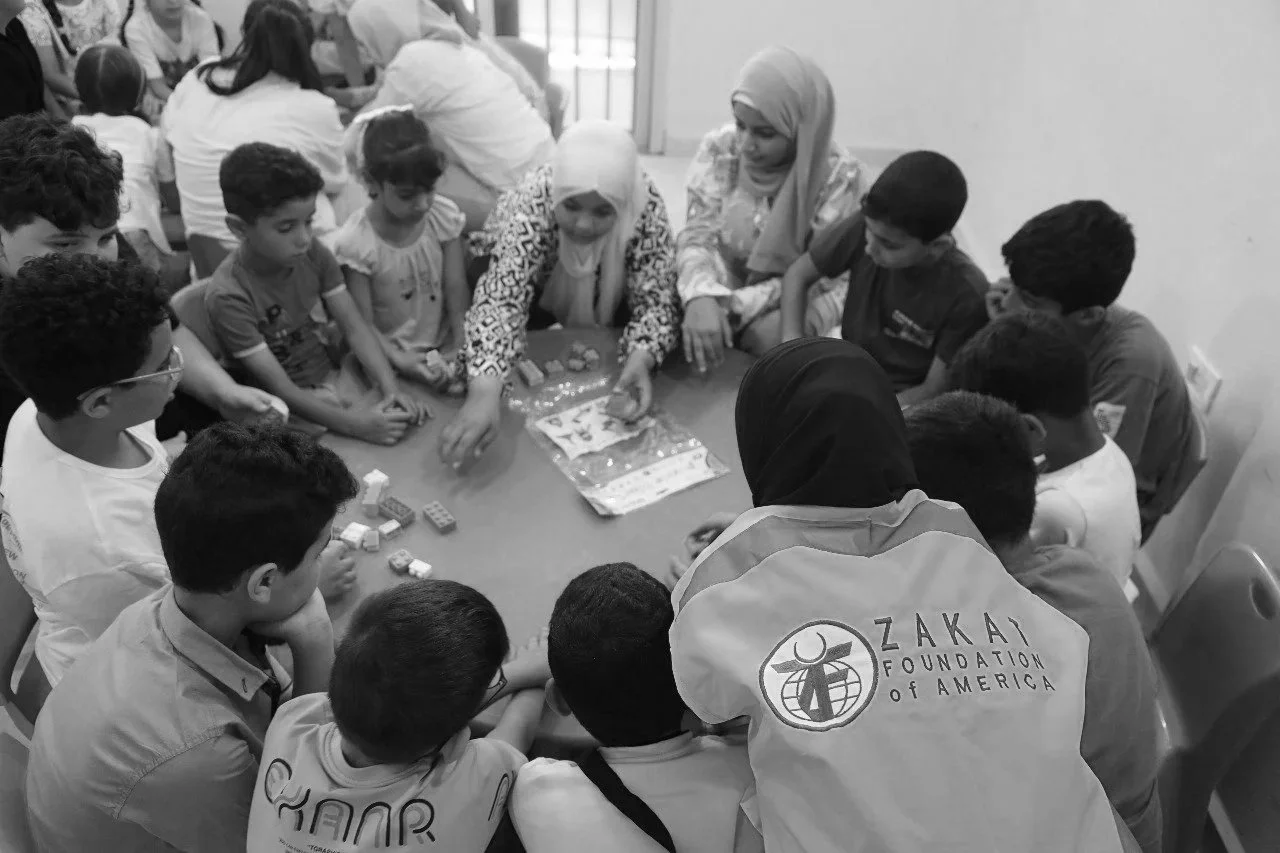 Women and children gathered around a table engaging in an activity, with some playing with small blocks. The woman on the right is wearing a hijab, and the woman on the left wears a patterned top. The back of a person's jacket has a logo and text for Zakat Foundation of America.