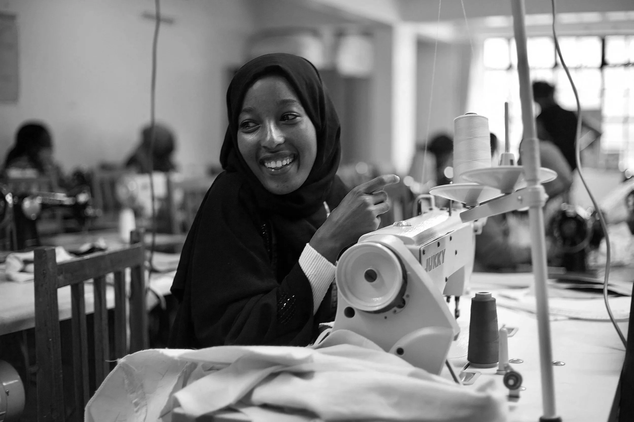 A young woman in a hijab smiling and pointing while working on a sewing machine in a busy sewing workshop.