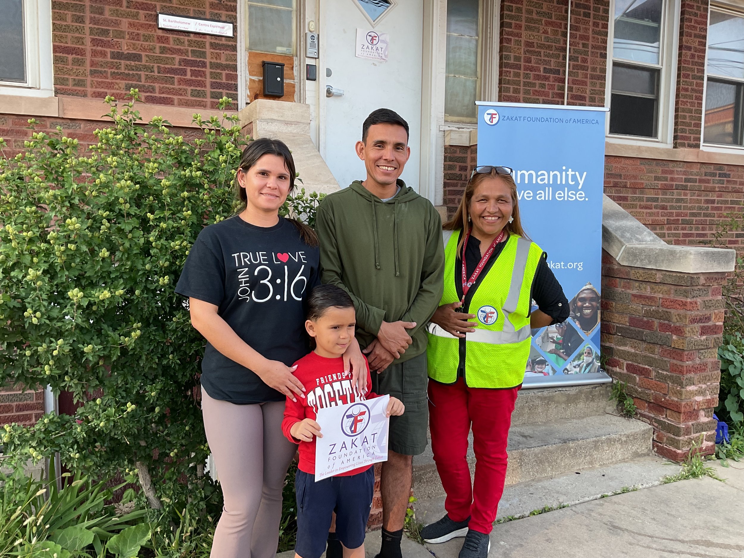 Group of four people standing in front of a brick building, two women, one man, and one children, holding a sign for Zakat Foundation of America, smiling for the camera. There is a greenery bush and a blue Zakat Foundation banner behind them.