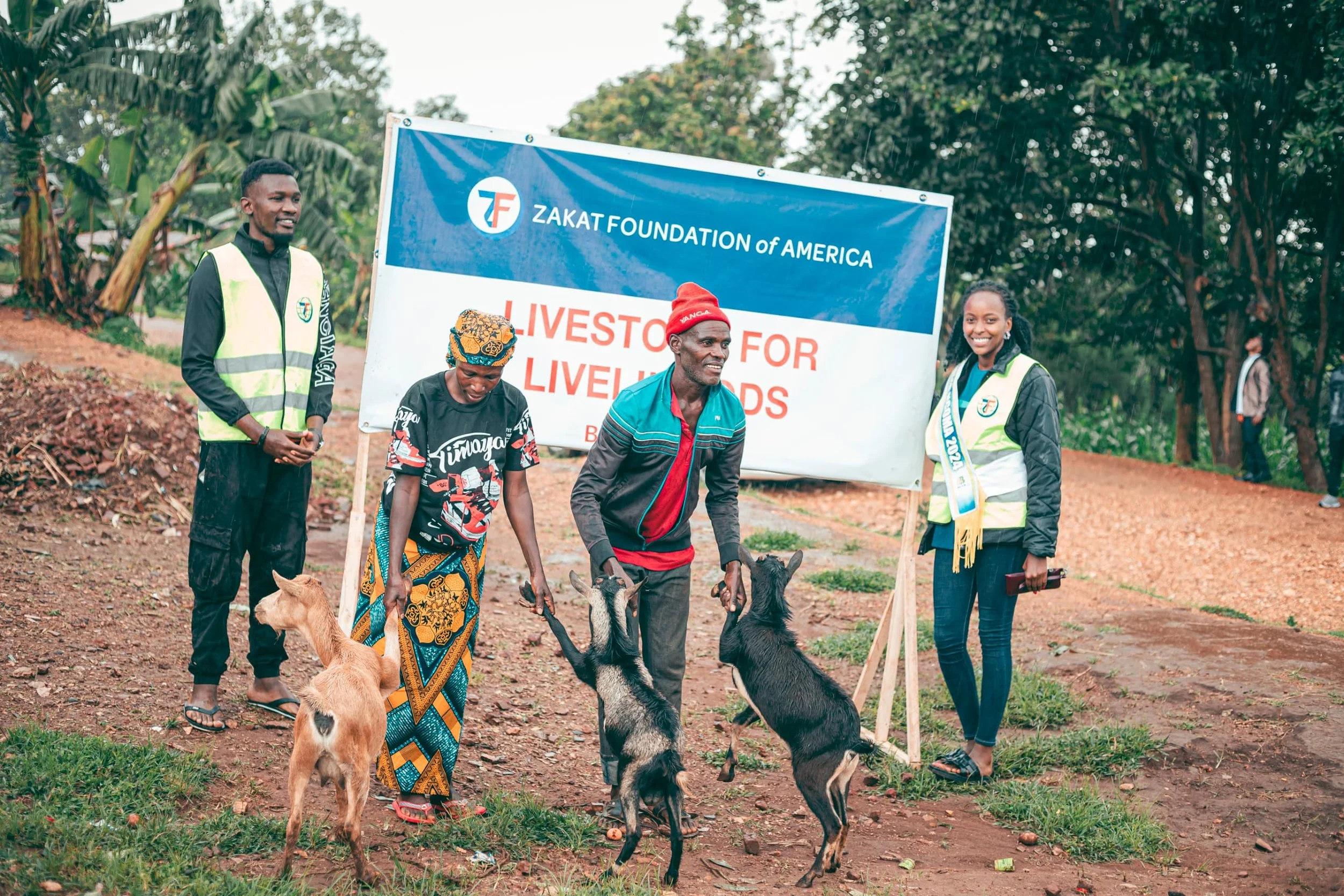 People at a community event for Zakat Foundation of America, with a sign that reads "Livestock for Livelihoods". Three individuals are engaging in a livestock donation, with goats present, and others are standing nearby.