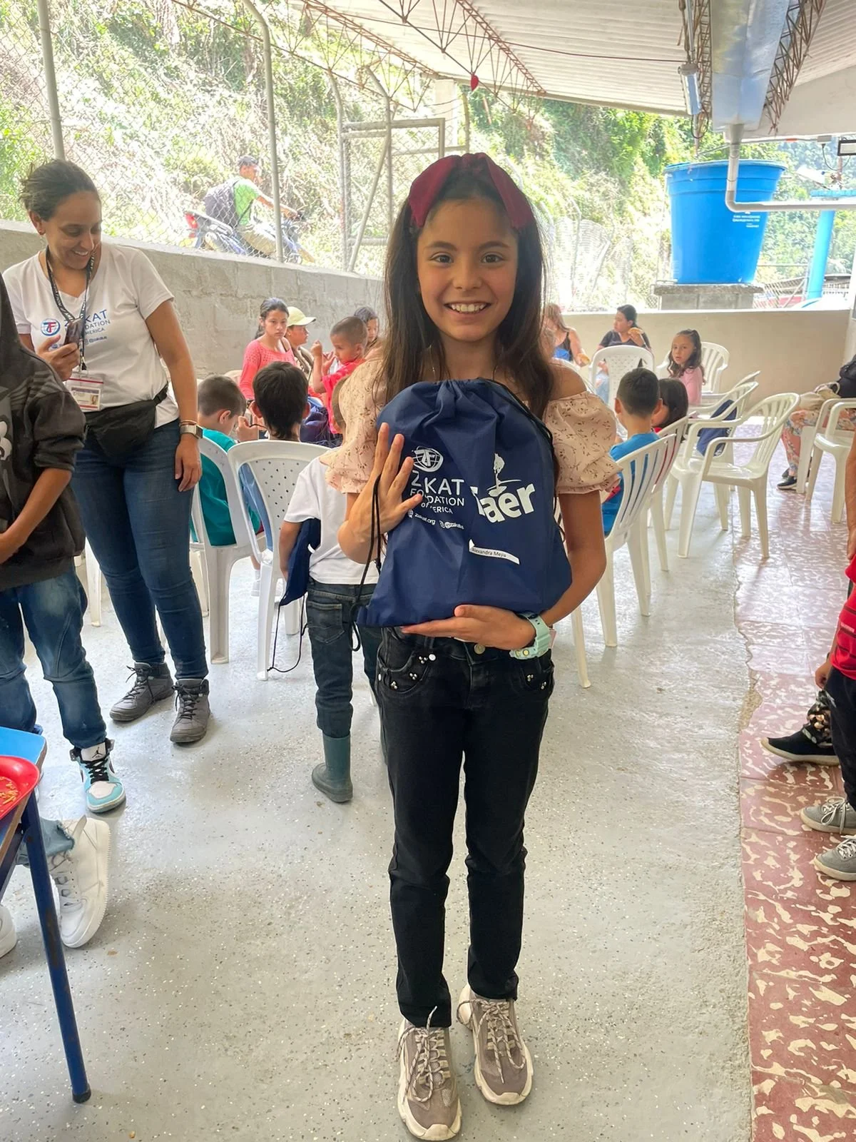 A young girl with long dark hair and a red headband holds a blue gift bag at an indoor event, smiling at the camera. There are children and adults seated on white plastic chairs in the background.