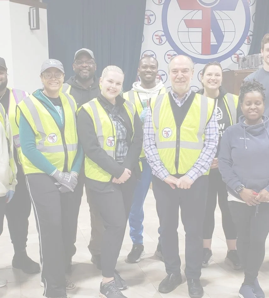 Group of diverse people in high-visibility vests and casual clothing standing together indoors in front of a background with the Zakat Foundation of America logo, smiling at the camera.