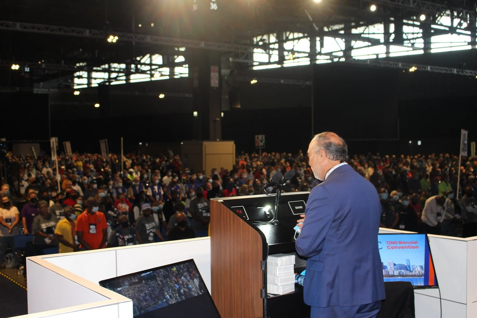Halil Demir in a blue suit addressing a large crowd at a convention, viewed from behind on a stage with monitors and microphones, inside a spacious indoor venue with high ceilings.