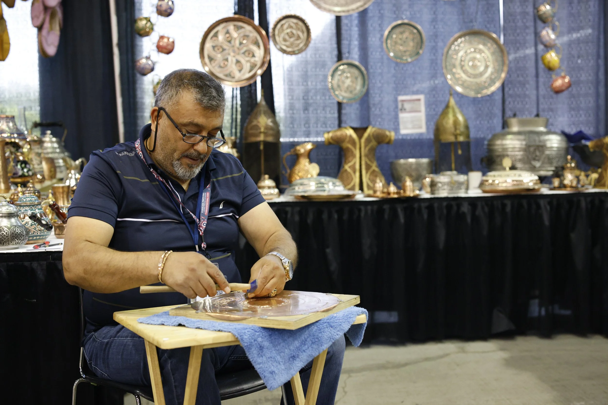 A man with glasses and a beard sitting at a small wooden table, working on a piece of metal with a hammer and chisel at an art or craft booth, with decorative metal items displayed in the background.