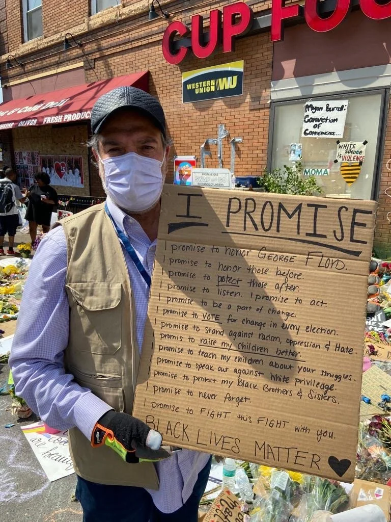 Halil Demir wearing a face mask and beige vest standing in a street holding a large cardboard sign with handwritten promises related to social justice and equality. Behind him are flowers and signs at George Floyd memorial site.