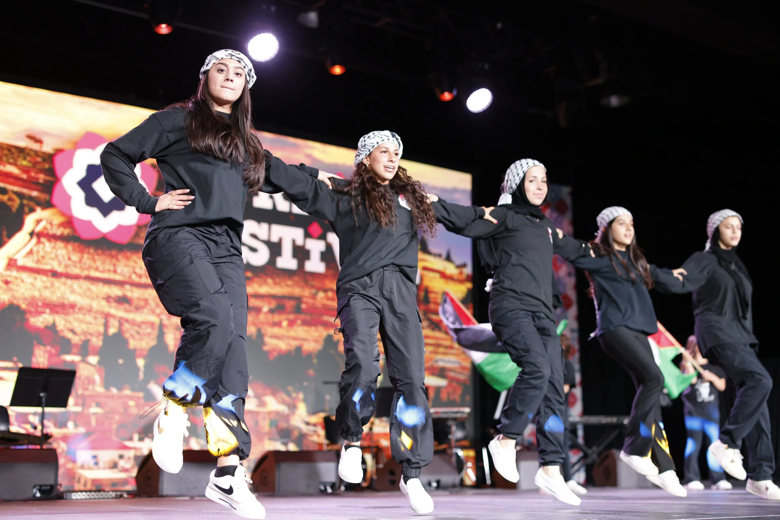 Five young women in black outfits and white bandanas perform a dance on stage, with a colorful scenic backdrop behind them.