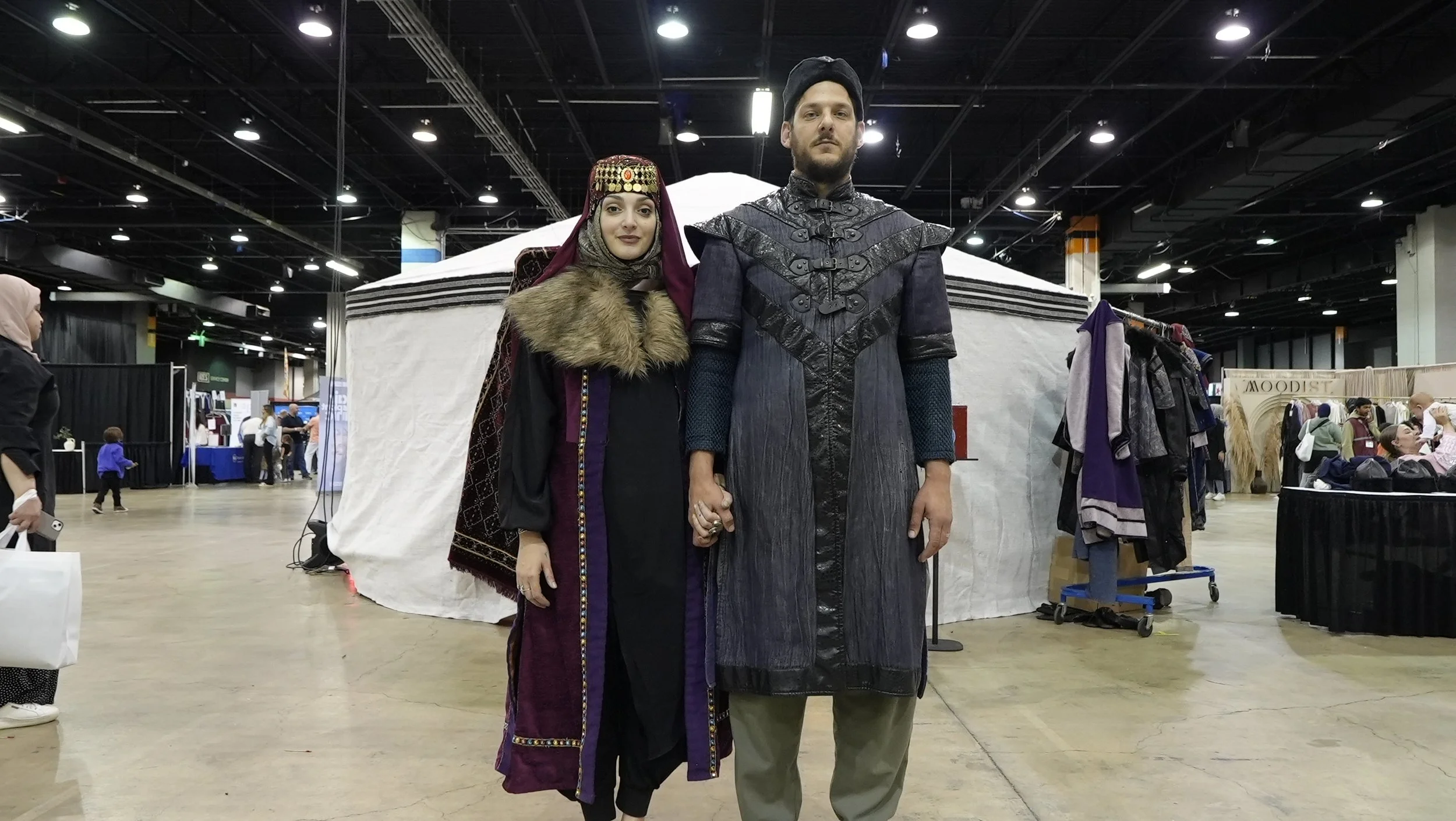A man and woman dressed in medieval or fantasy costumes, holding hands, standing in an indoor market or convention center.