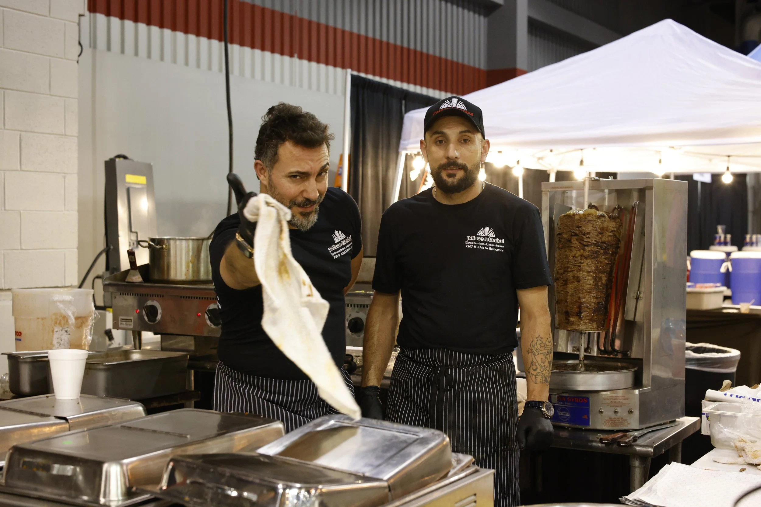 Two male chefs standing in a food stall kitchen, one holding a cloth or towel, with a vertical rotisserie with meat in the background and various kitchen equipment around.