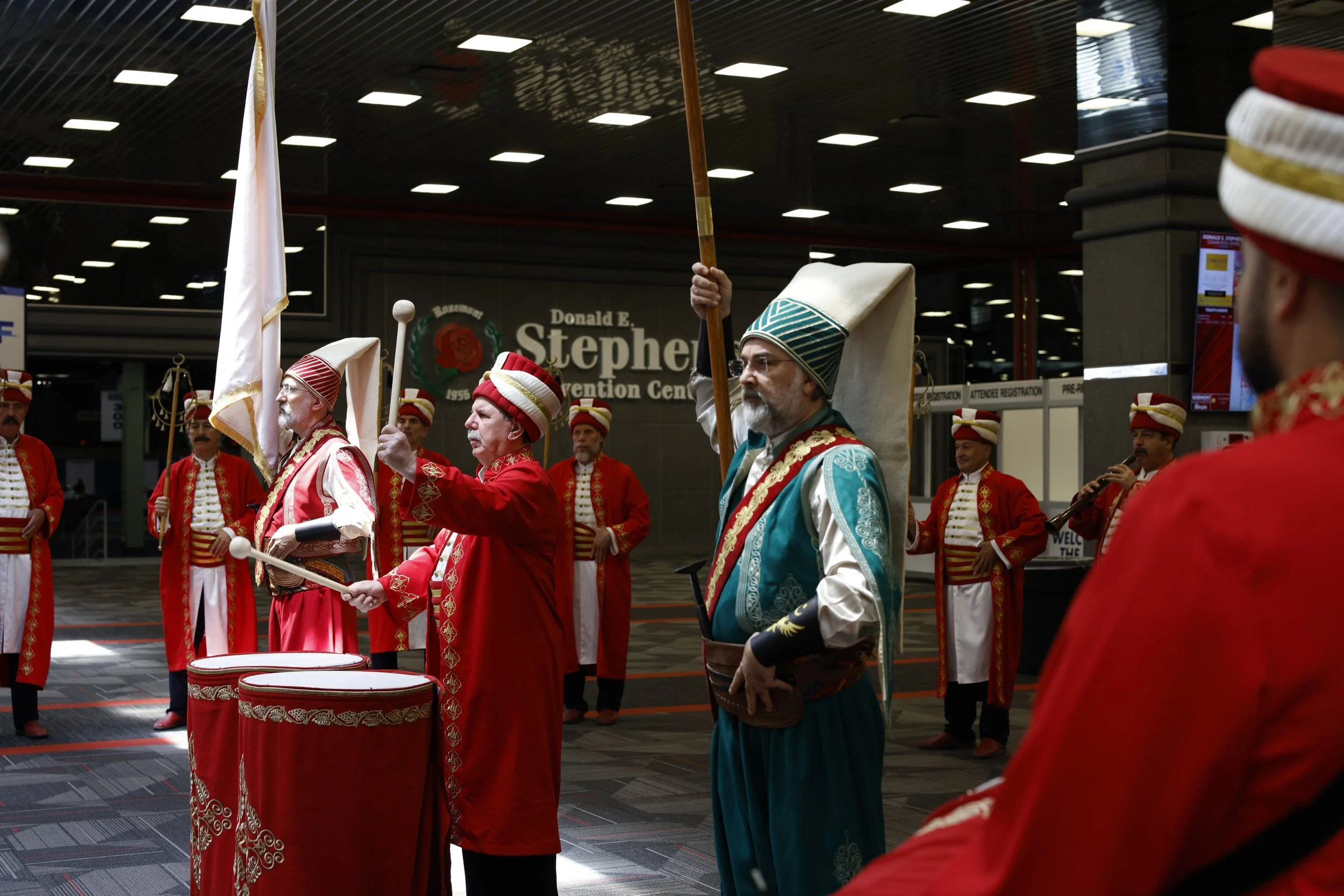 A group of men dressed in colorful traditional Ottoman-style costumes, some playing drums and others holding flags, are performing at the Donald E. Stephens Convention Center.