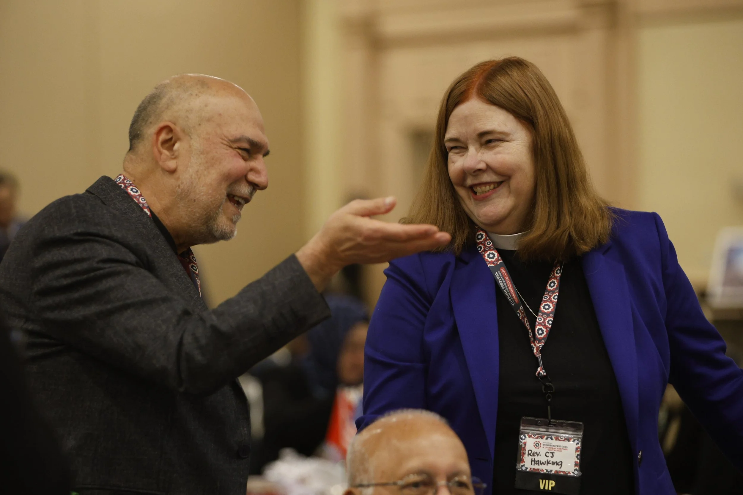 Two people are smiling and talking at a conference, one with a beard and gray hair, the other with red hair wearing a name tag that reads 'Rev. CJ Hawking'.