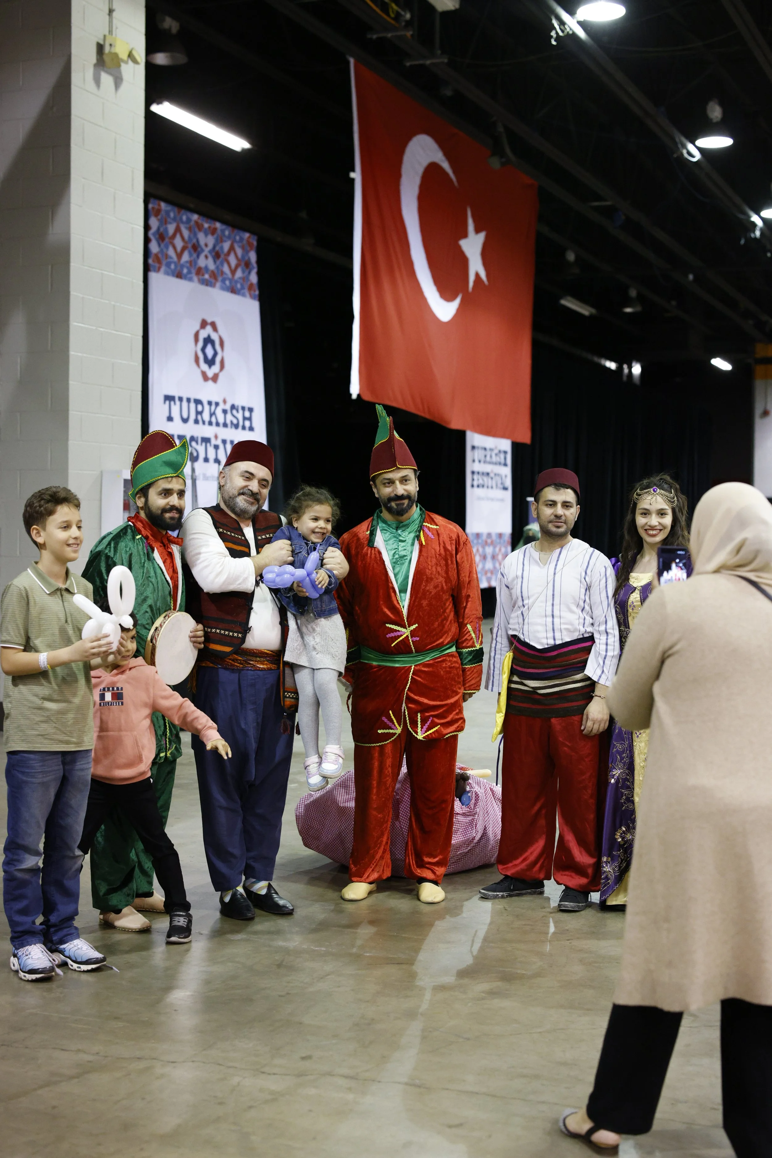 Group of people dressed in traditional Turkish and cultural costumes, posing for a photograph at an indoor cultural event with Turkish flags and banners in the background, including one with the Turkish national flag.