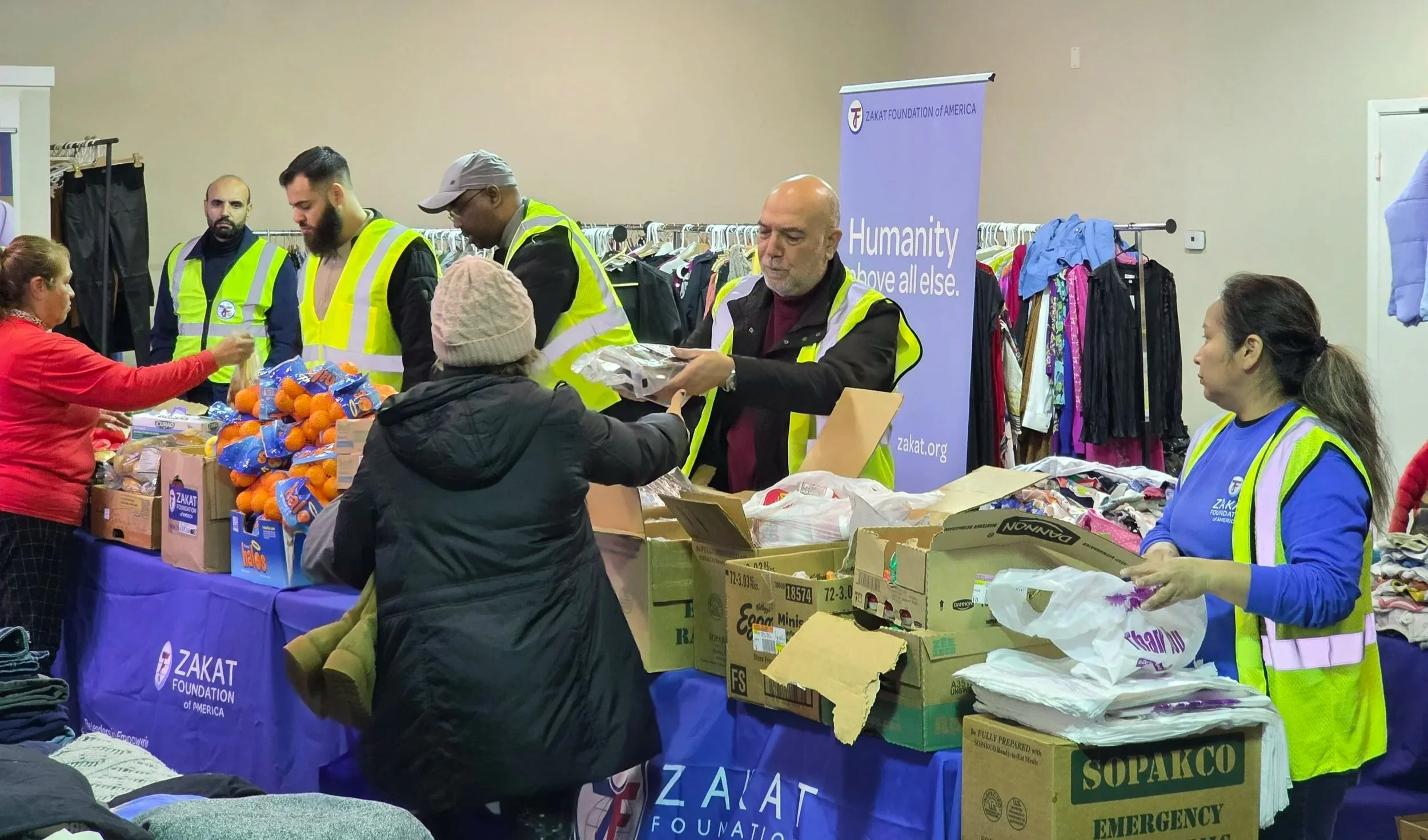 Volunteers at a Zakat Foundation of America event distributing food and supplies to people in need. There are clothing racks and boxes of food on the tables, and volunteers are wearing yellow safety vests.