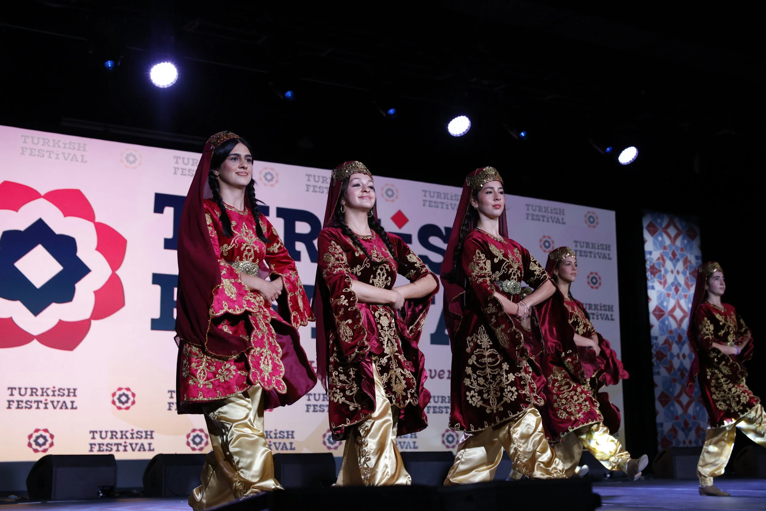 Six women dressed in traditional Turkish costumes, performing a dance on stage at a Turkish Festival, with a backdrop displaying a logo and the words "TURKISH FESTIVAL."