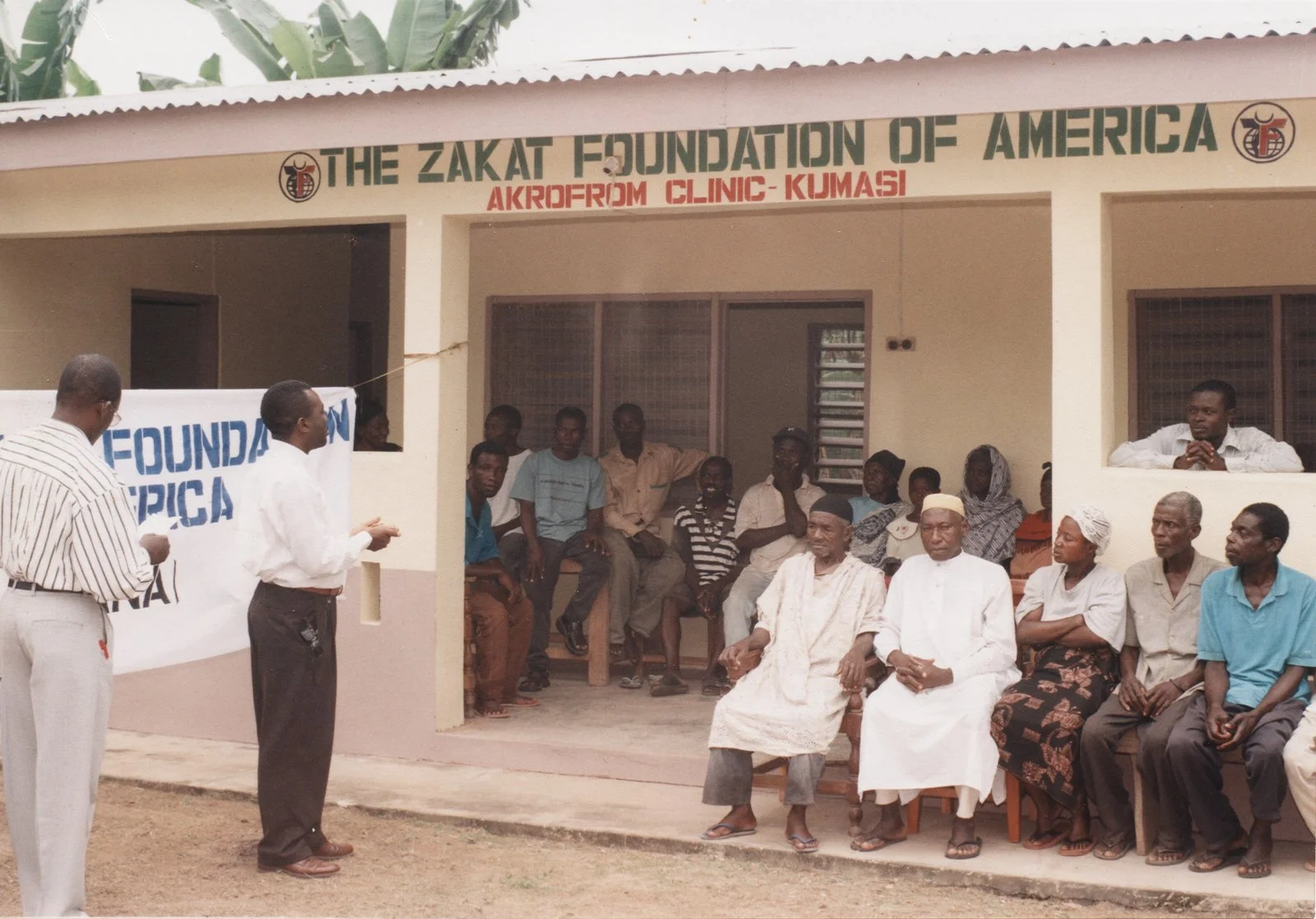 Community gathering outside the Zakat Foundation of America building in Kumasi, Ghana, with people sitting on chairs and listening to speakers.