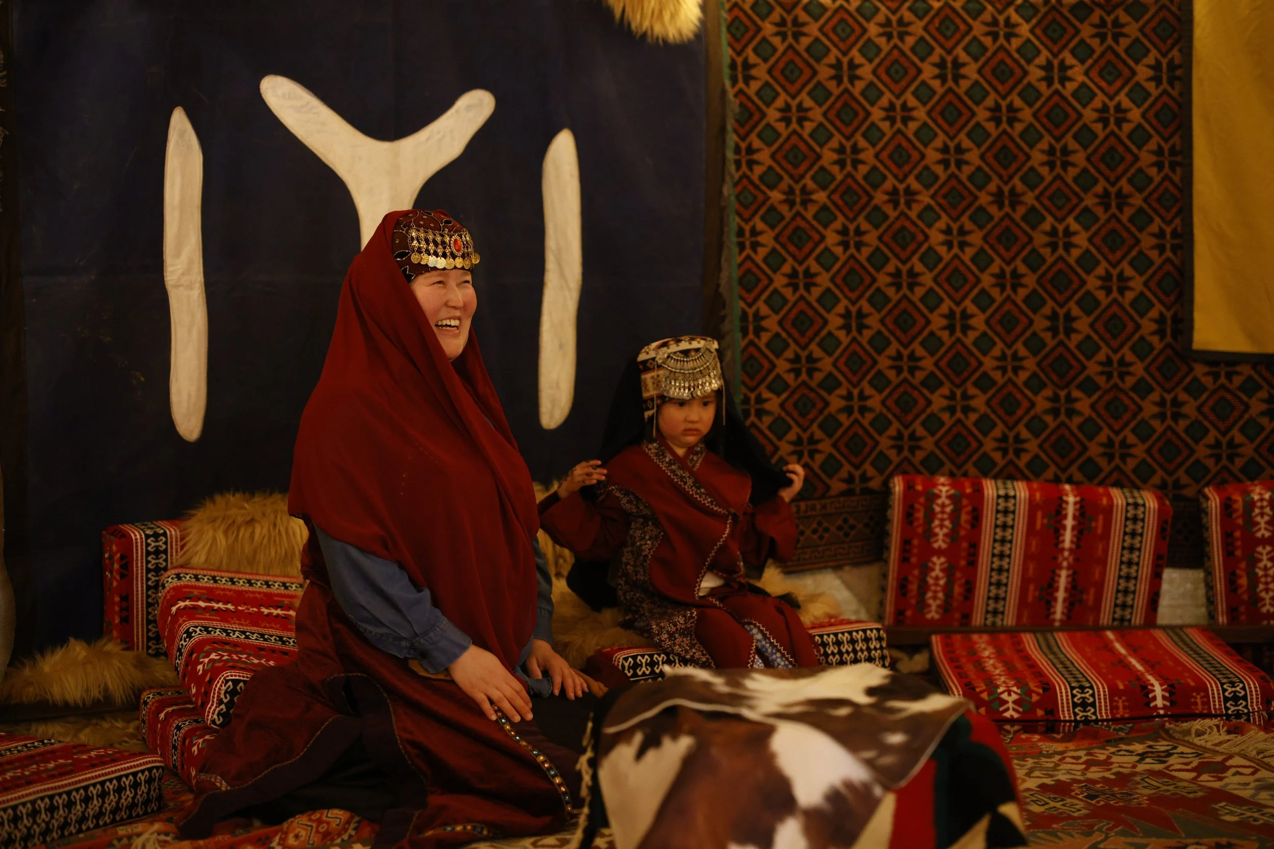A woman and a child dressed in traditional Central Asian clothing, sitting on a patterned rug in a decorated room with textiles and artwork.