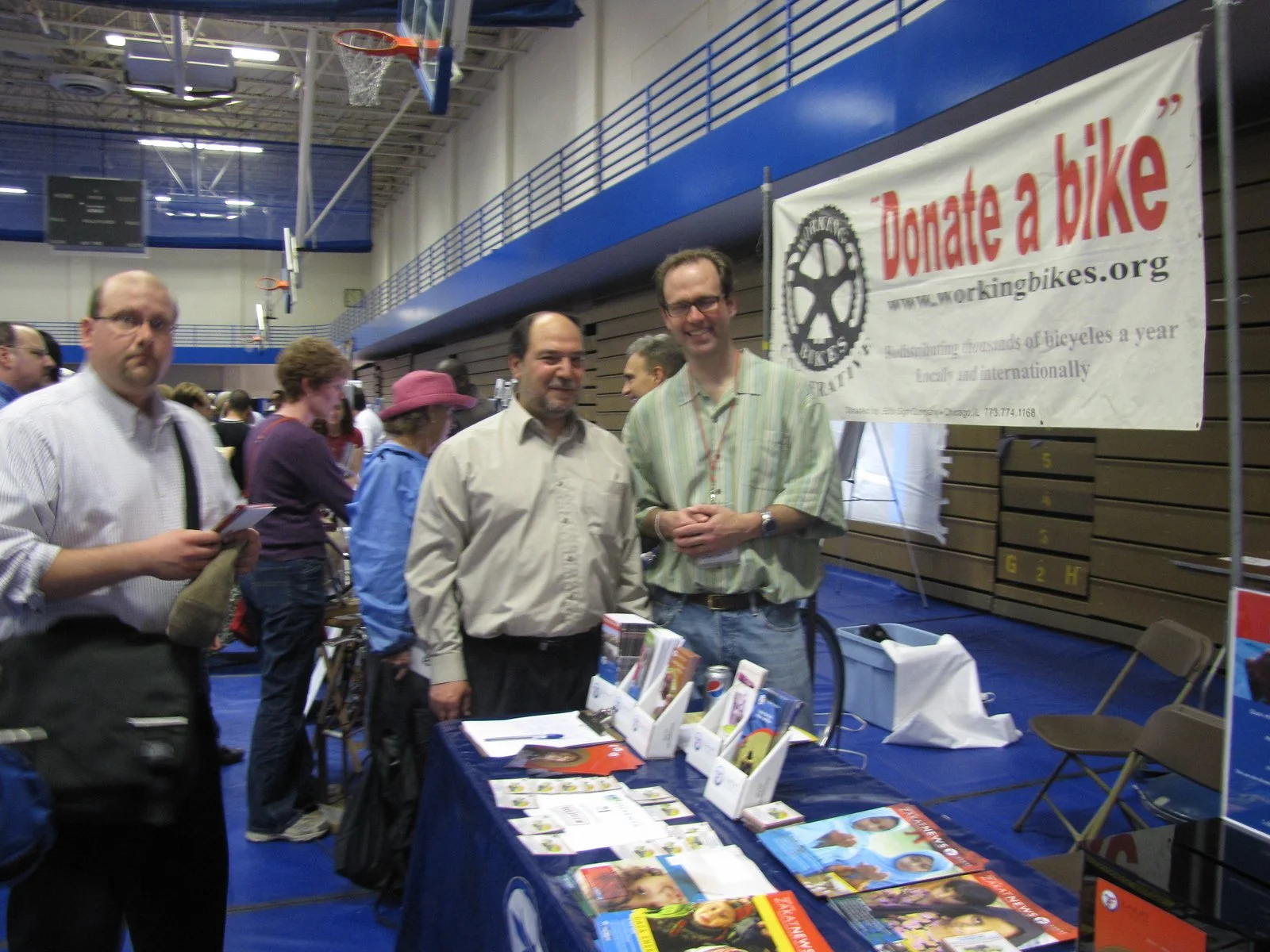 People gathered at a booth promoting bike donations at an indoor event. Two men are standing behind a table displaying brochures and informational materials, with a large banner in the background that says 'Donate a bike' and includes a website URL.