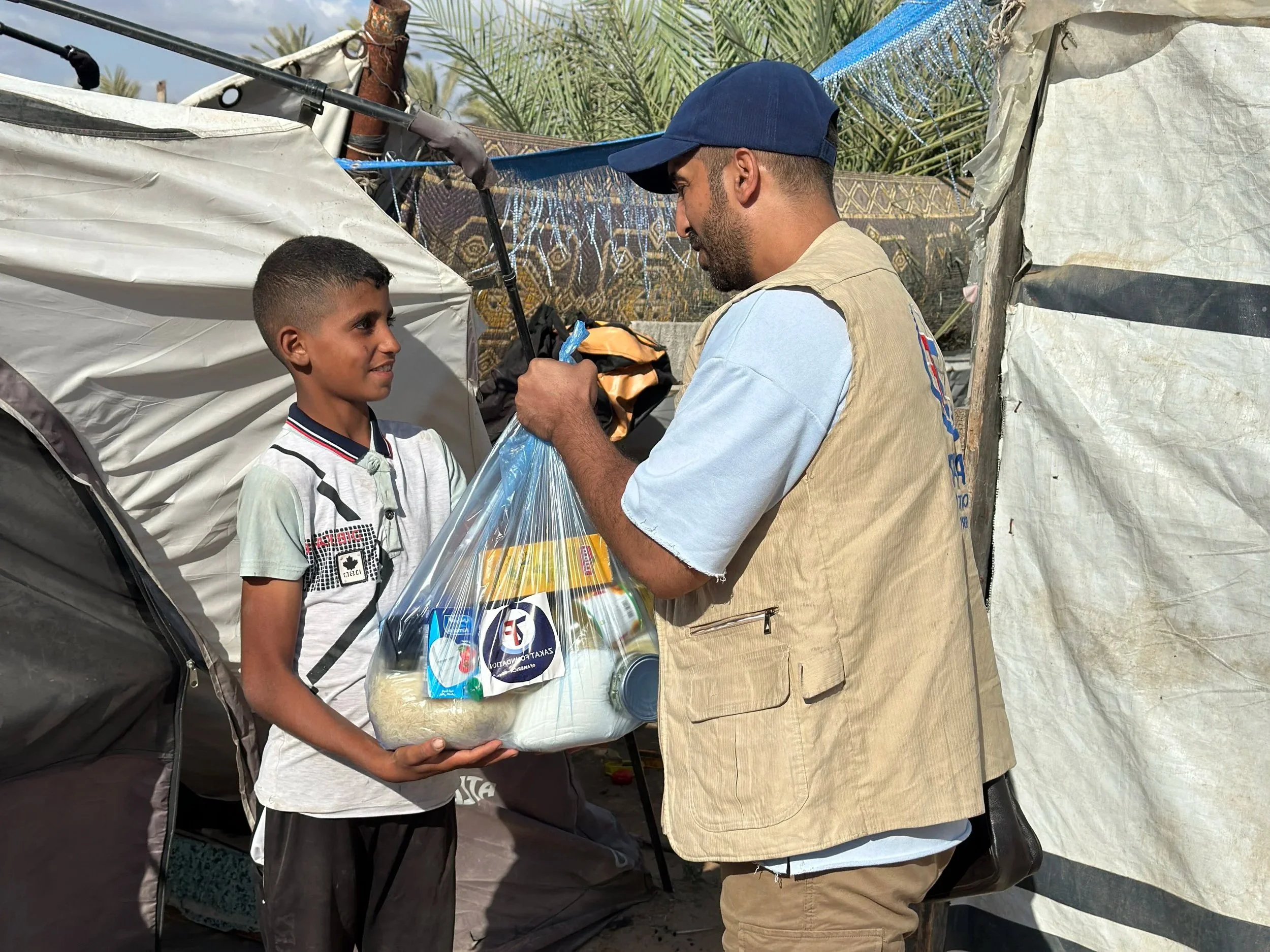 A boy receiving a bag of supplies from a man in a makeshift outdoor setting with tents and palm trees in the background.
