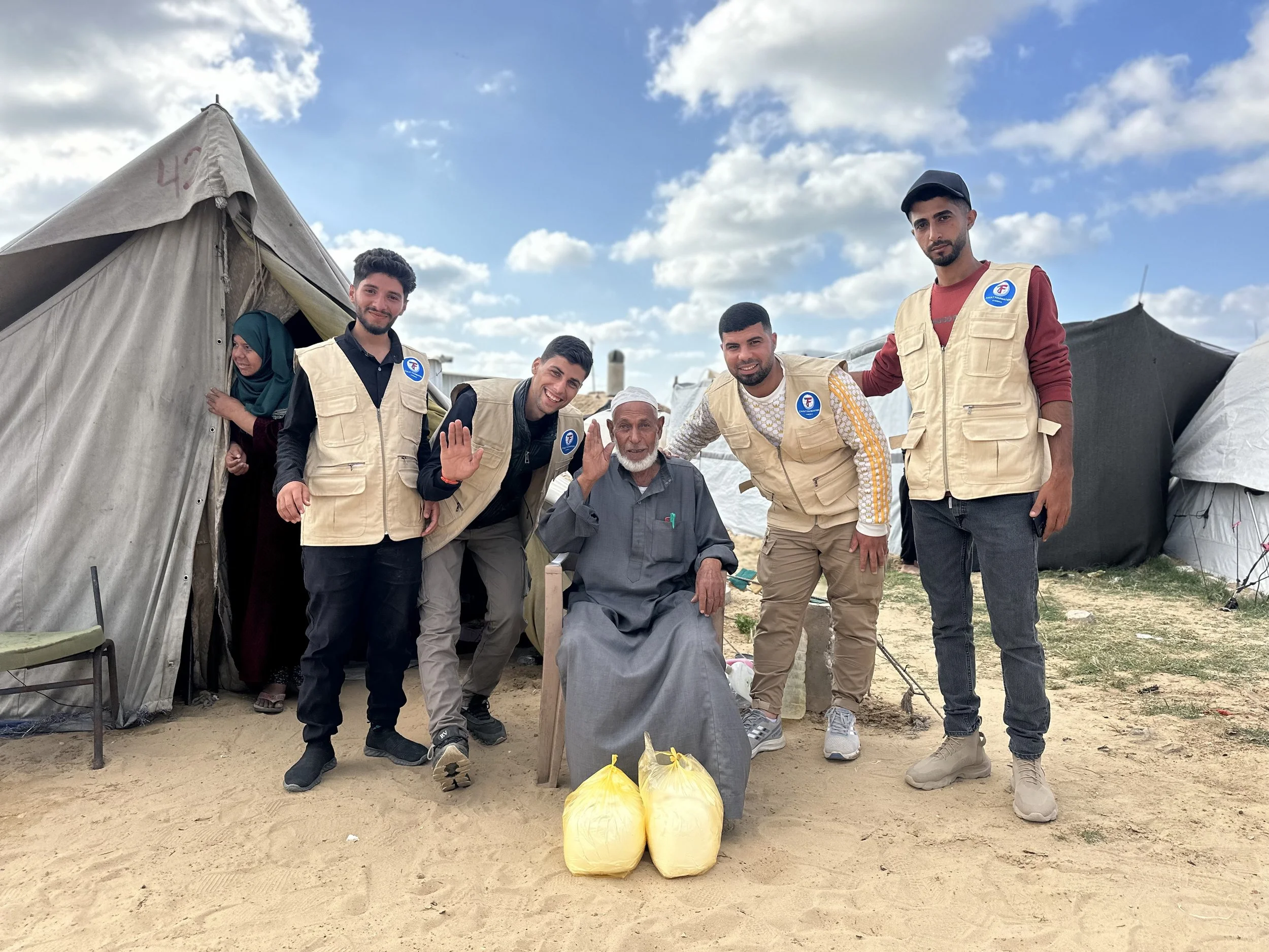 Group of five young volunteers wearing beige vests standing with an elderly man in a refugee camp with tents in the background. The elderly man is seated, smiling, waving, and has two yellow bags placed in front of him. The scene is outdoors under a partly cloudy sky.