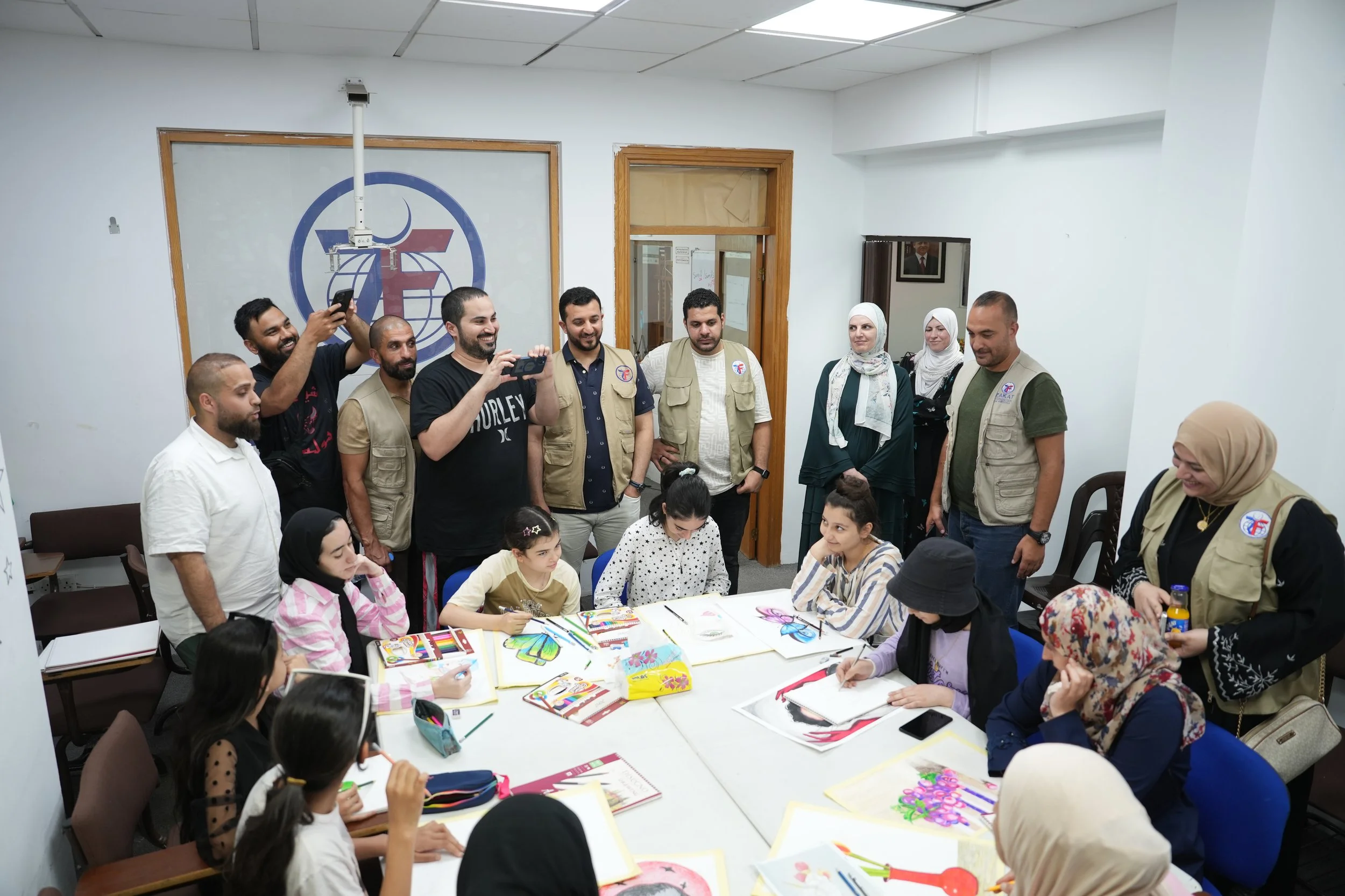 Children sitting at a table drawing and coloring, surrounded by adults standing and taking photos in a bright room with the Zakat Foundation of America logo on the wall.