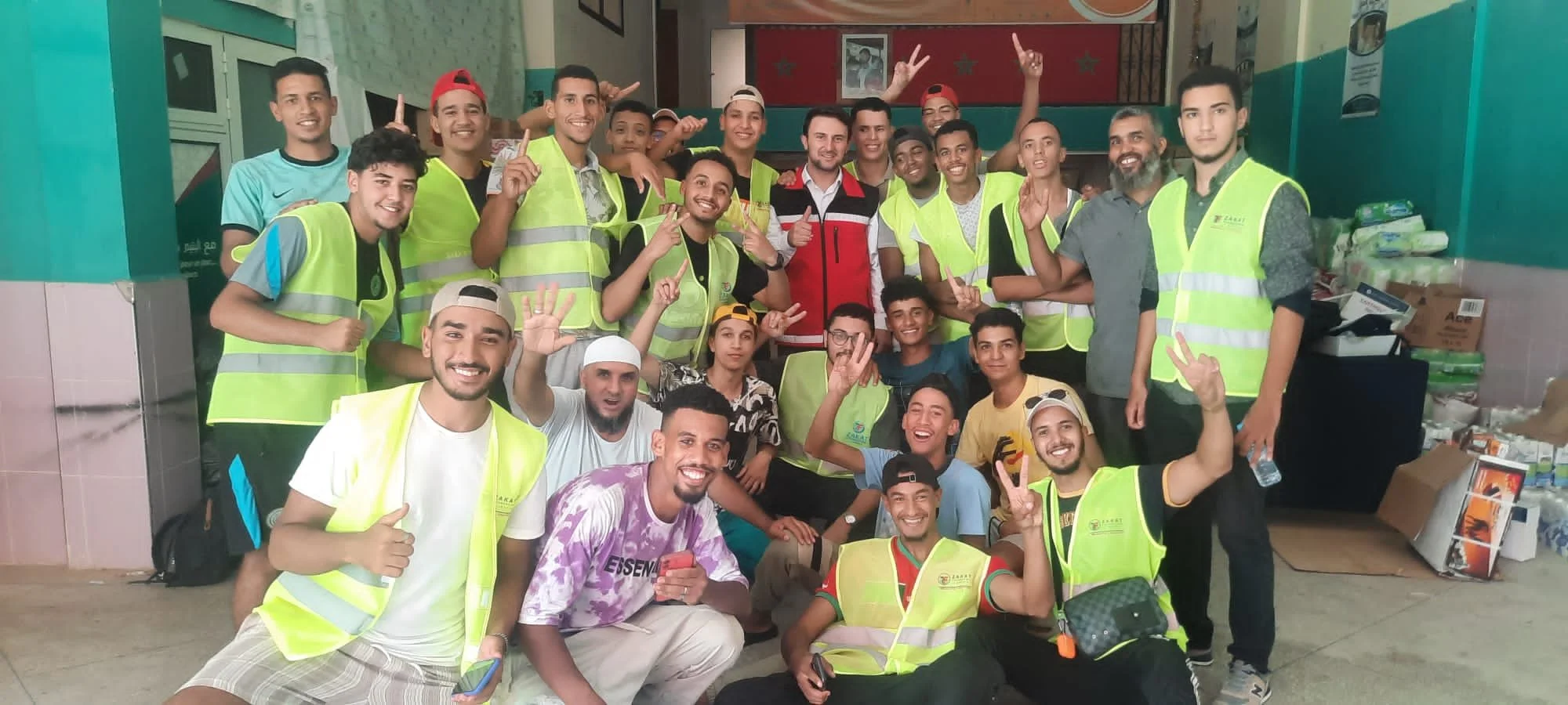 Group of smiling young people, many wearing yellow safety vests, gathered in an indoor space with some people making peace signs. Background includes boxes and supplies, indicating a community or volunteer event.