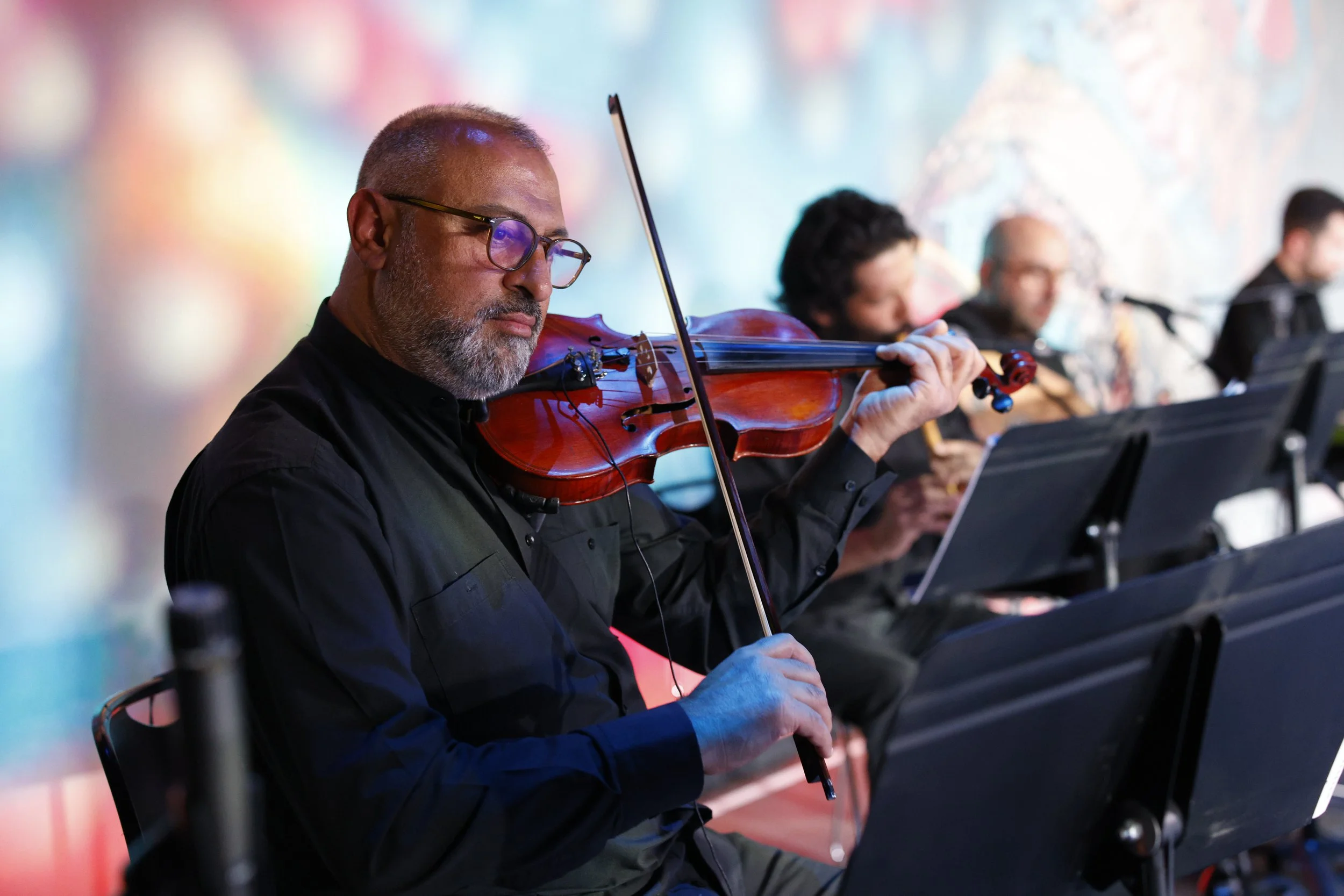 A man with glasses playing a violin during a musical performance, with other musicians in the background.