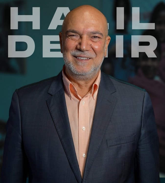 Halil Demir smiling and  wearing a dark suit and light orange shirt, standing in front of a blue background with large gray text that reads 'HALIL DEMIR'.