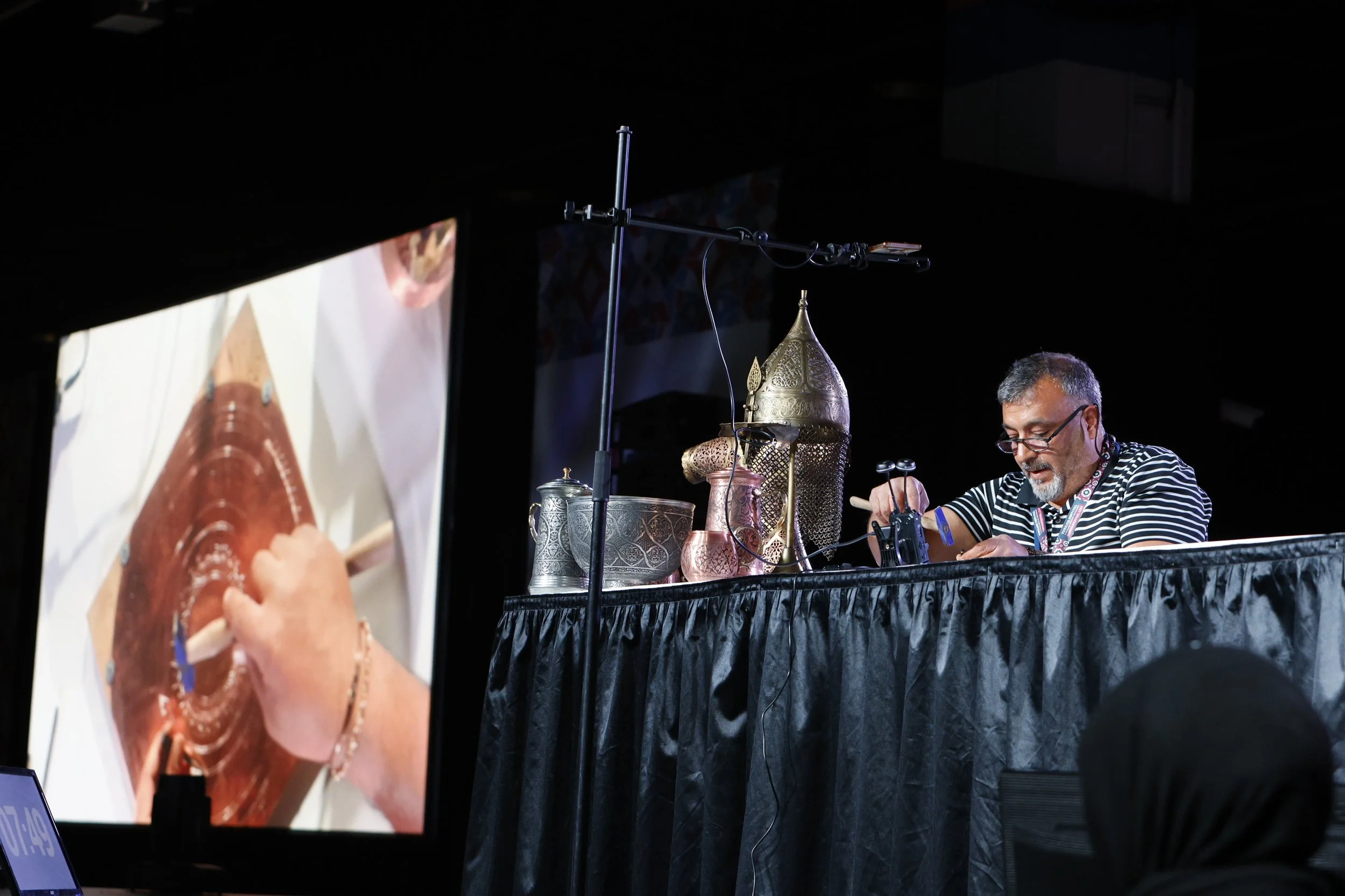 A person sitting at a table with traditional metal and brass decorative items, operating a pottery wheel or craft device, with a large screen showing a close-up of their hands working on a clay piece.