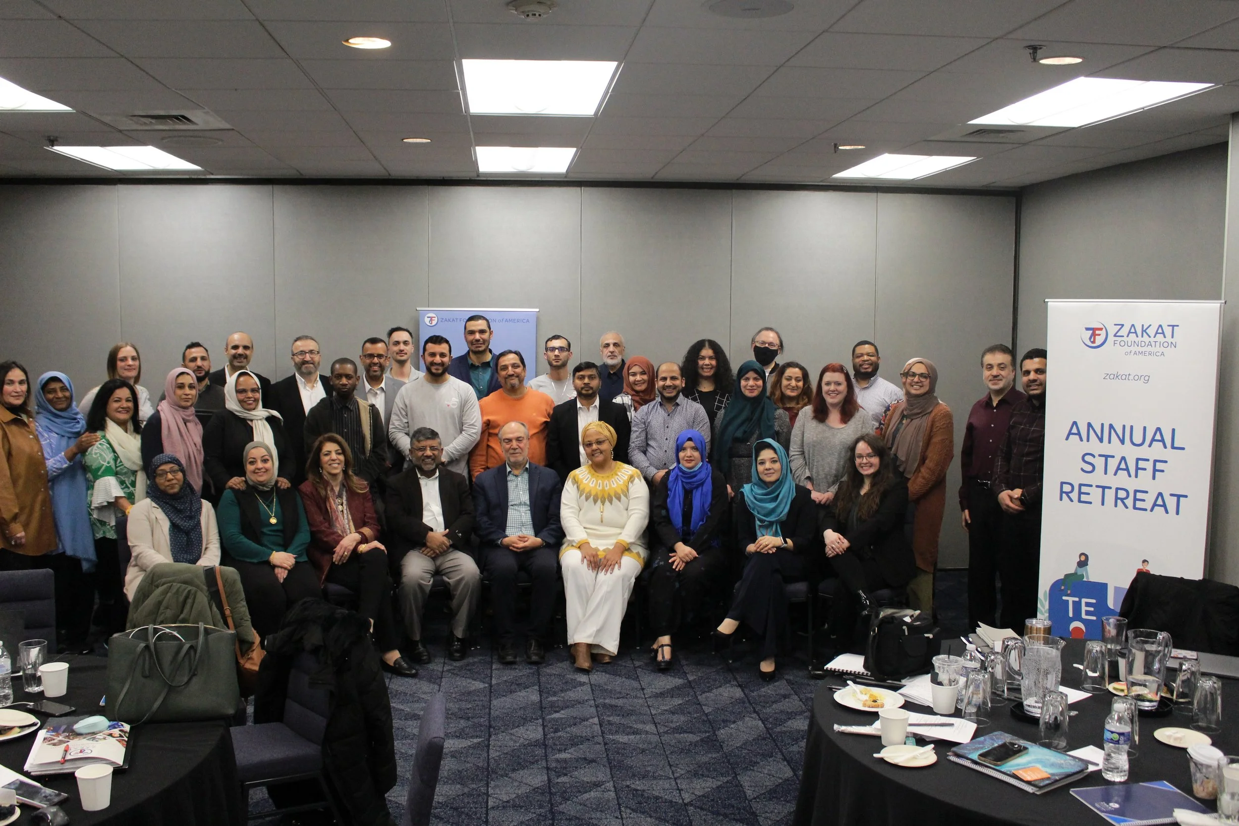 A group of diverse people attending an annual staff retreat, posing for a group photo in a conference room with tables and a banner that reads 'Zakat Foundation of America'.