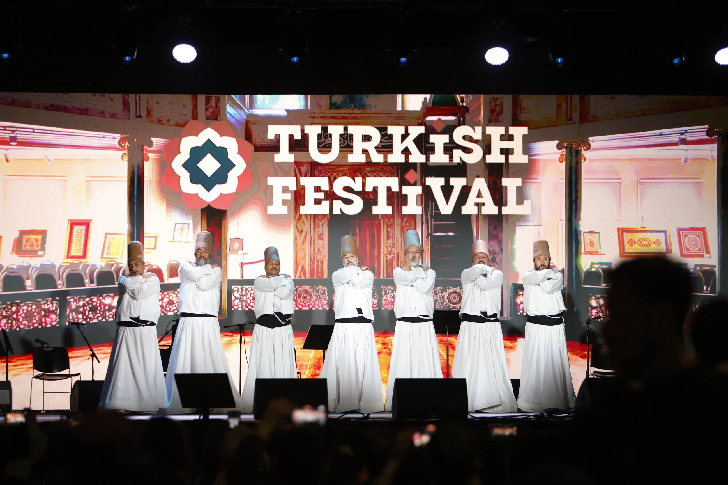 Men dressed in traditional Turkish costumes performing a dance on stage at the Turkish Festival, with a backdrop featuring Turkish decor and the event name displayed prominently.