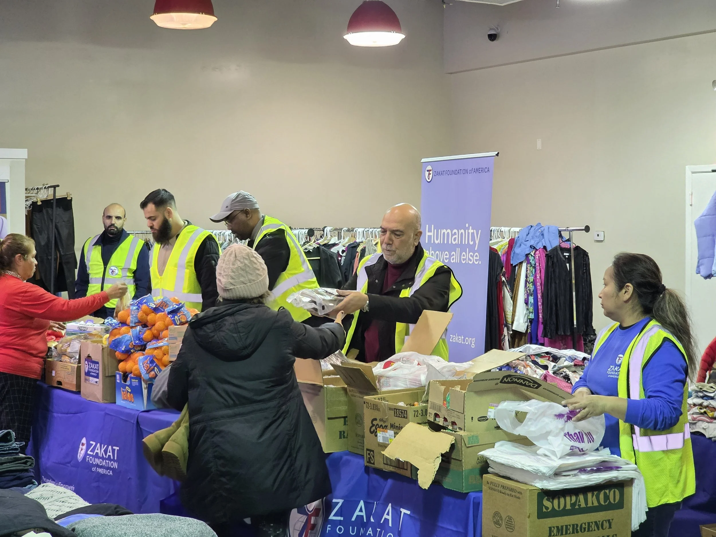 People wearing yellow safety vests distributing food and clothing at a charity event organized by Zakat Foundation of America, with tables of supplies and clothes in the background.