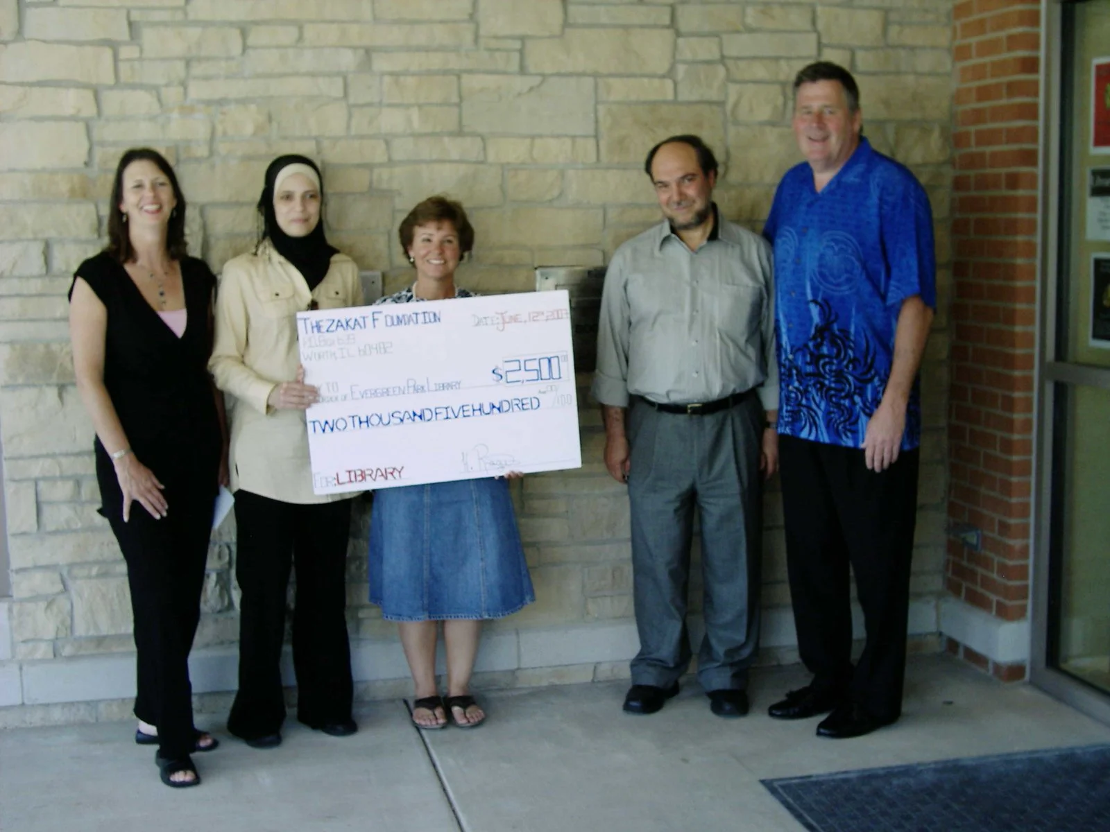 Six people standing outside, holding a large check for $2,500 made out to the Evergreen Park Library, celebrating a donation. The group includes five adults, all smiling, with a brick and stone wall behind them.