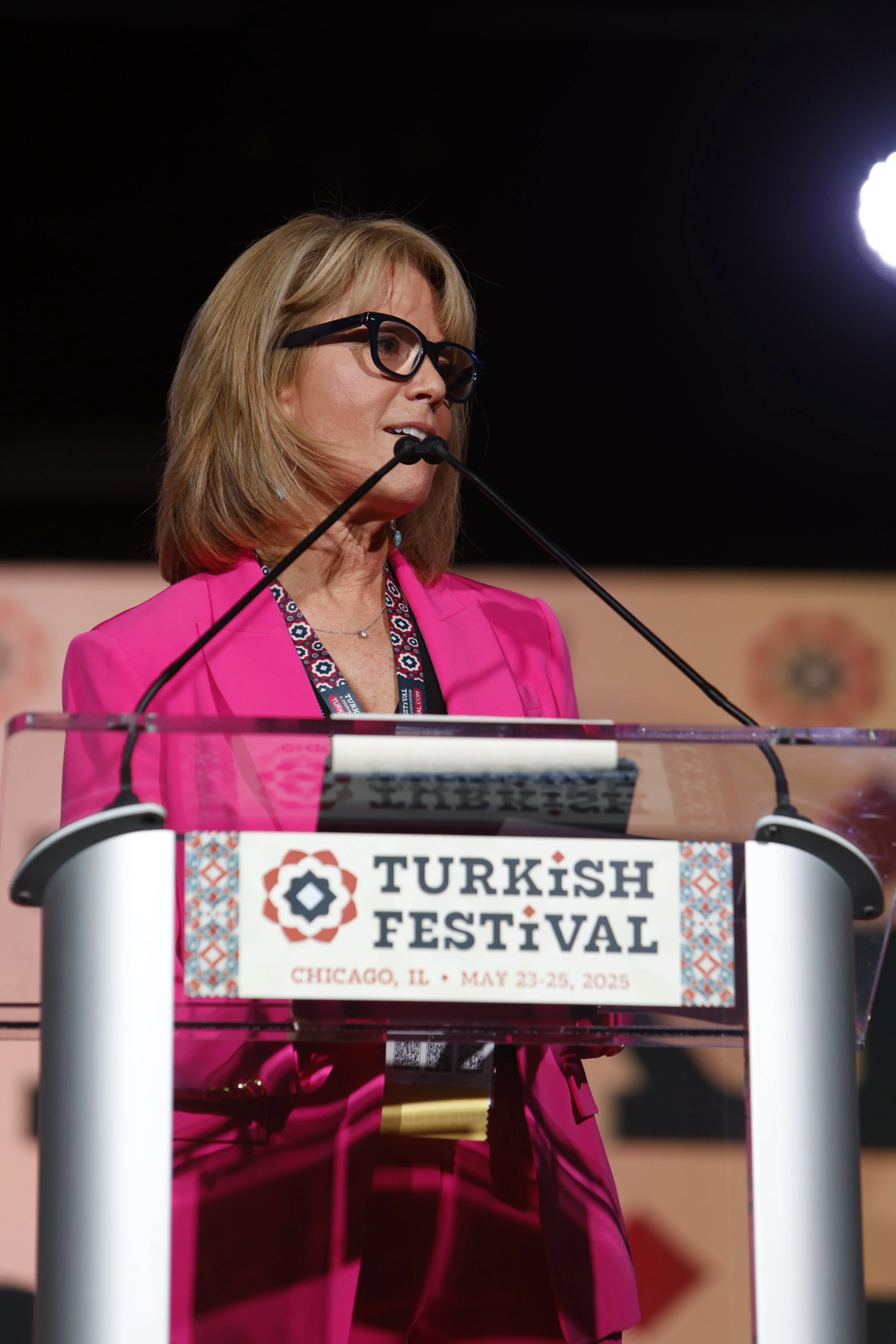 A woman speaking at a podium during the Turkish Festival in Chicago, Illinois, held from May 23 to 25, 2025. She is wearing a bright pink blazer, black glasses, and a patterned lanyard.