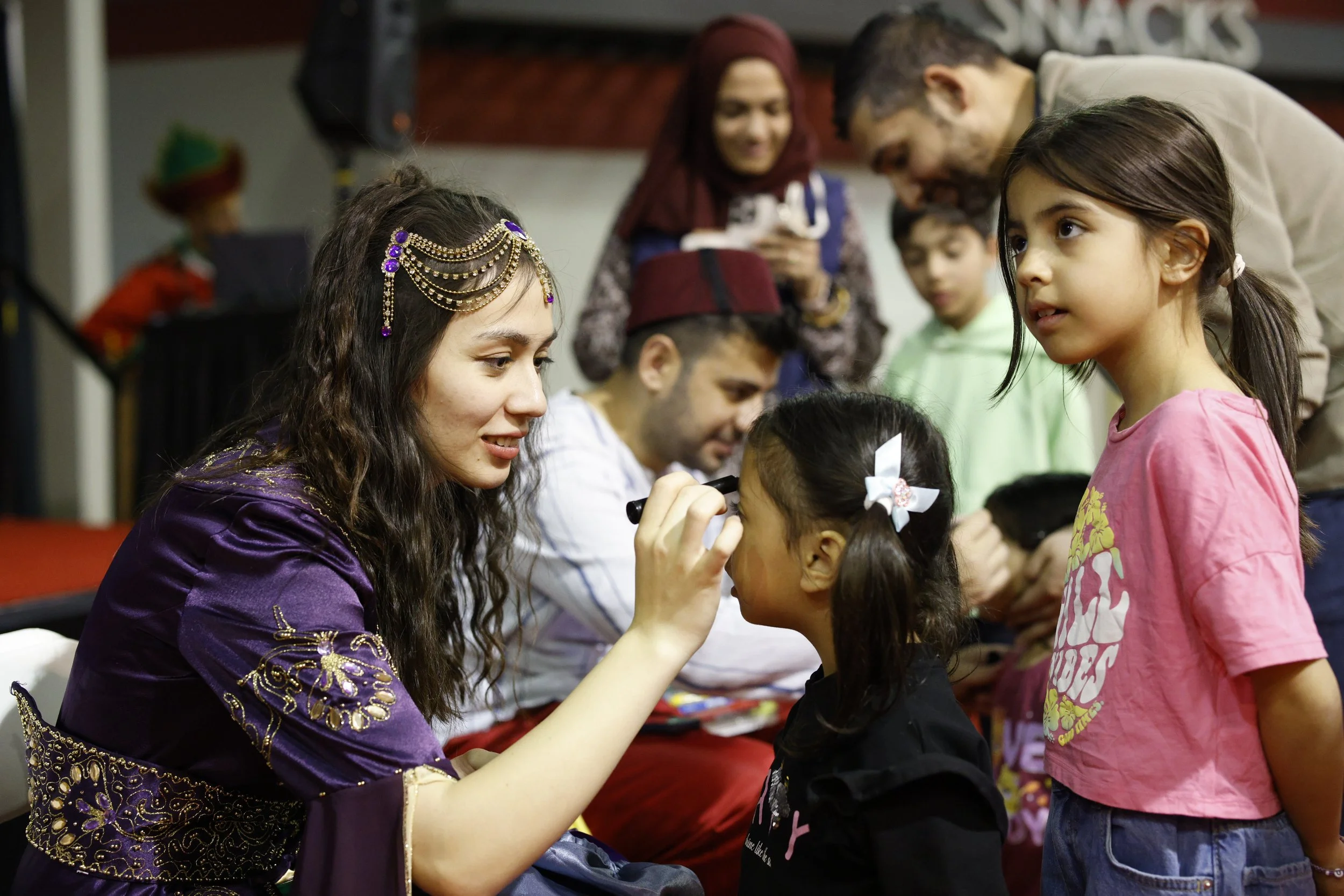 A woman dressed in traditional attire with jewelry is painting a young girl's face at a cultural event. Other children and adults are gathered around.”}