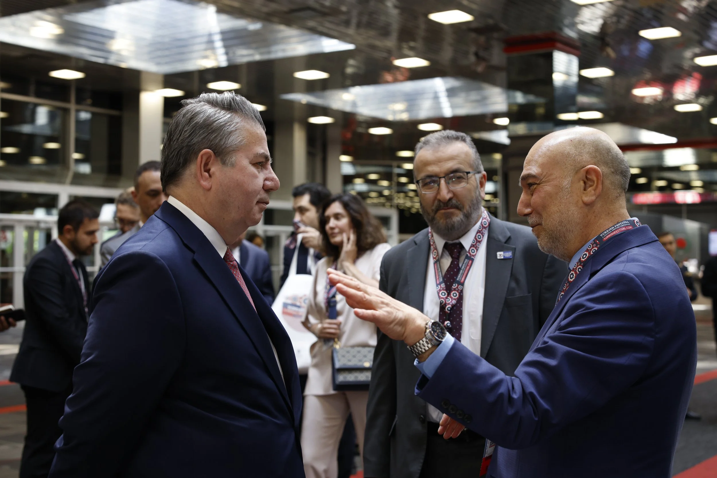 Three men in suits engaged in conversation at a professional event, with several other people in the background.