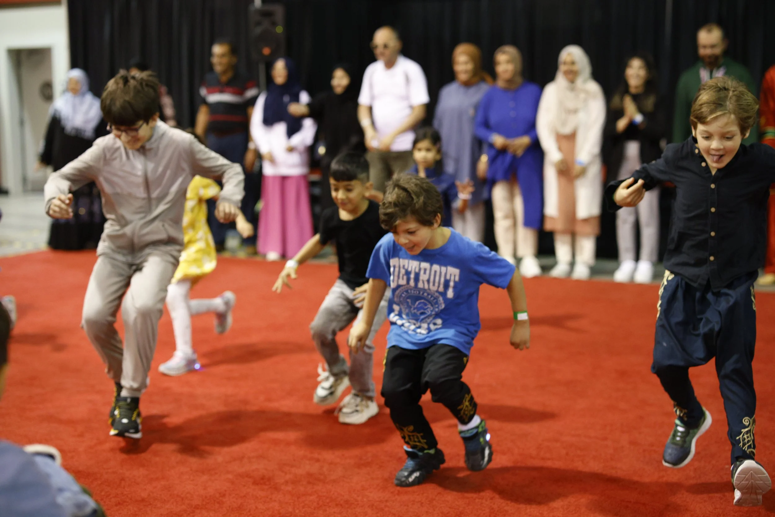 Children dancing or jumping on a red carpeted floor during an indoor event, with adults and various individuals standing in the background.