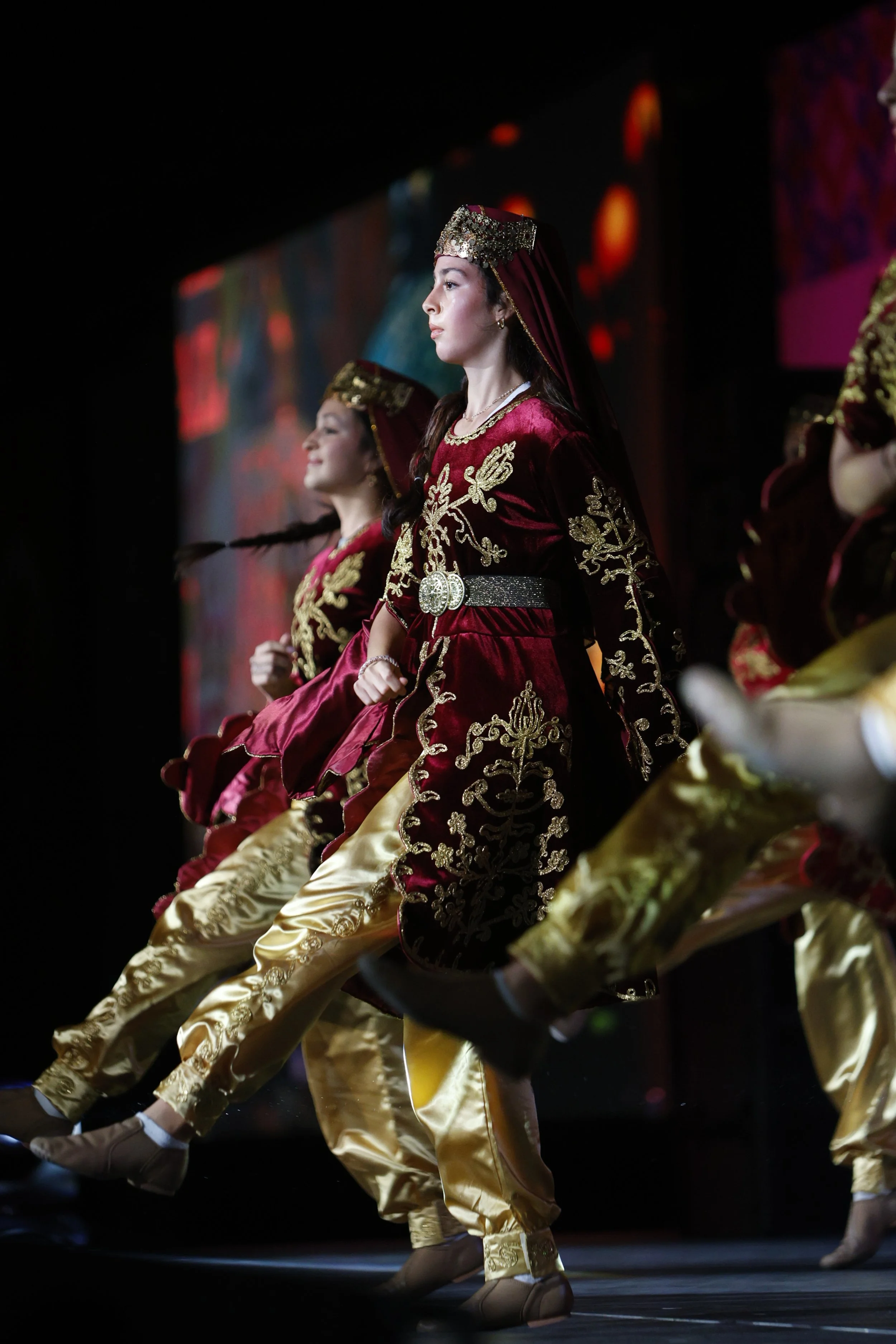 Women dressed in traditional costumes performing on stage with velvet tops and gold embroidered pants.
