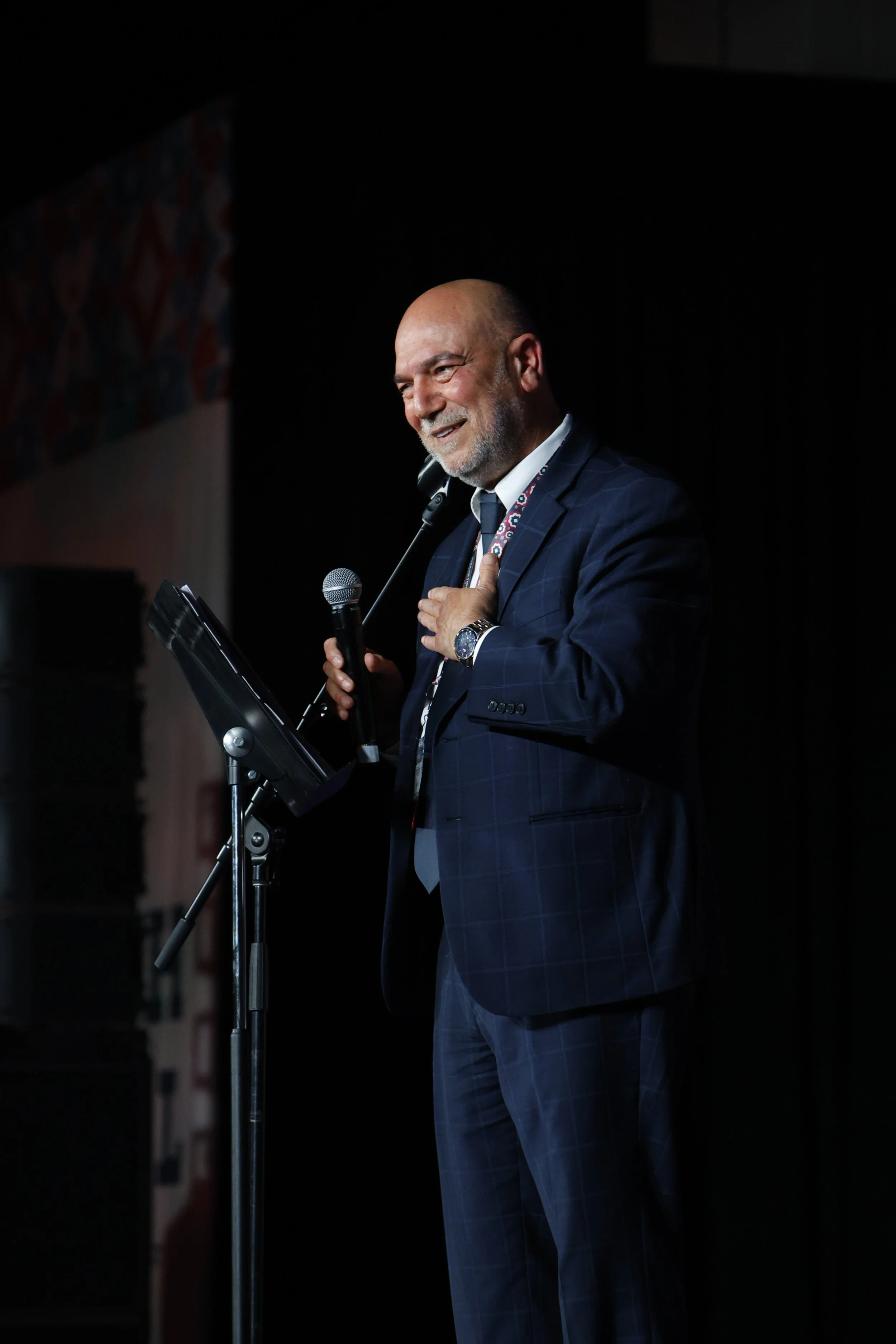 Halil Demir in a suit and tie speaking at a podium with a microphone, holding his hand over his heart, smiling, on a dark stage.