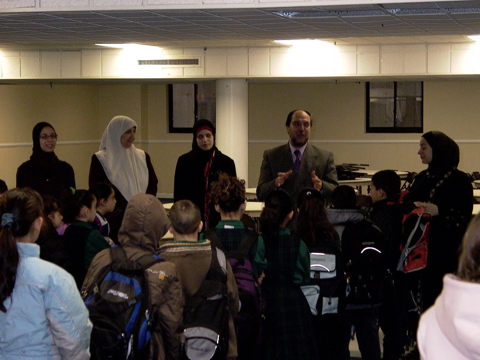 A group of children with backpacks listening to a man speaking, alongside four women, in an indoor room.