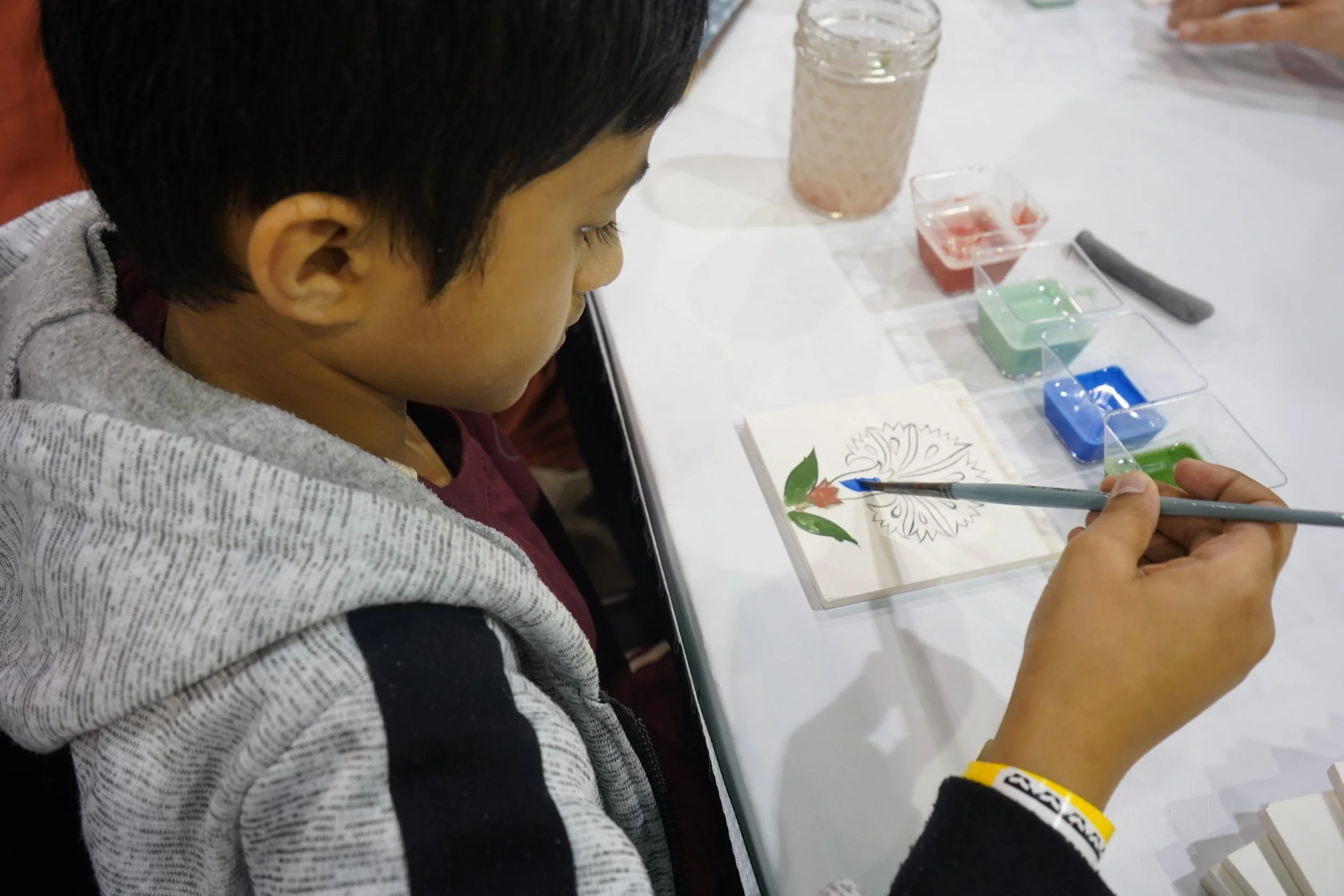 A young boy sitting at a table, painting a colorful flower on a book page with watercolor paints.